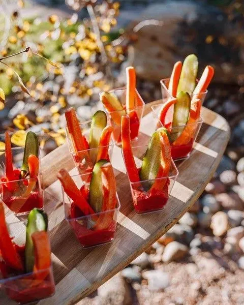 Vegetable sticks (carrots, cucumber) in small cups with red dip, arranged on a wooden board outdoors.
