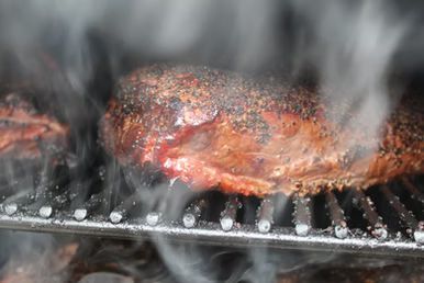 Smoked meat cooking on a grill, surrounded by smoke. The meat is dark and coated in seasoning.