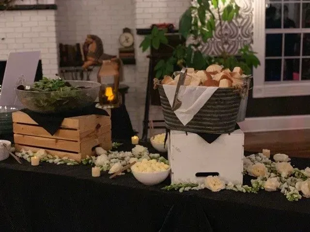Buffet table with salad, bread, and cheese, decorated with white flowers and greenery. The setting appears to be indoors with a brick wall backdrop.