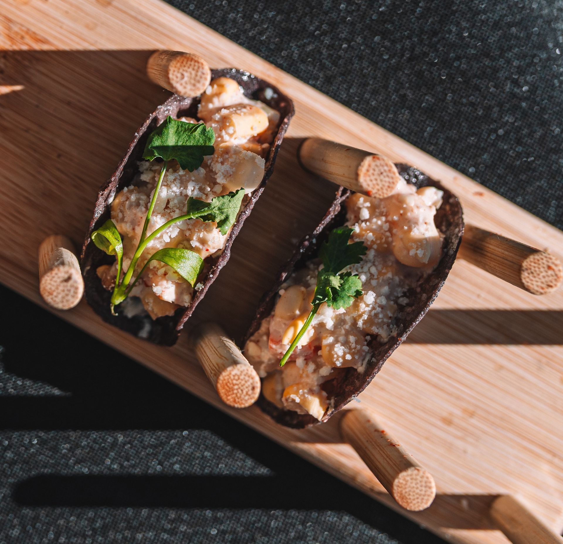 Two filled tacos on a wooden board with wooden accents. Light and dark colors, garnished with herbs.