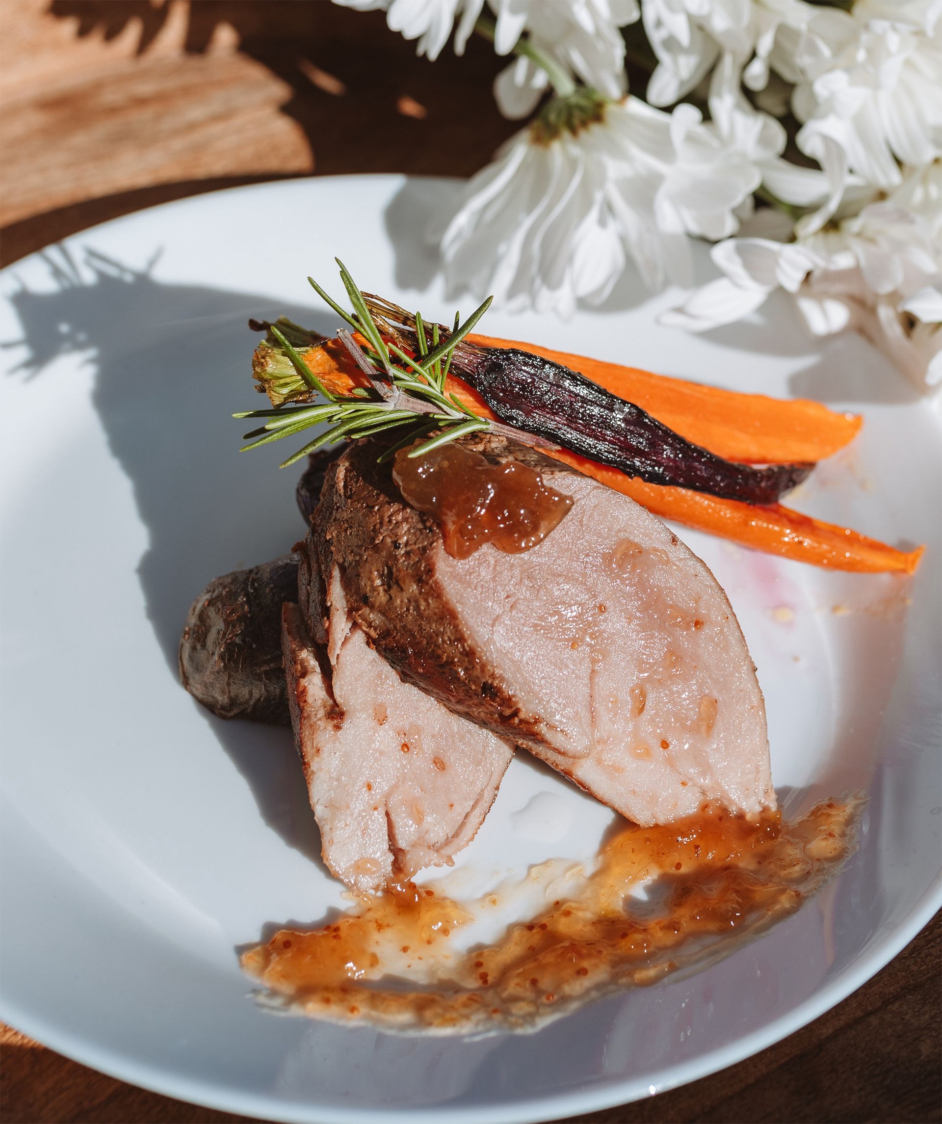 Plate of sliced meat with sauce, carrots, rosemary sprig, and white flowers in the background.