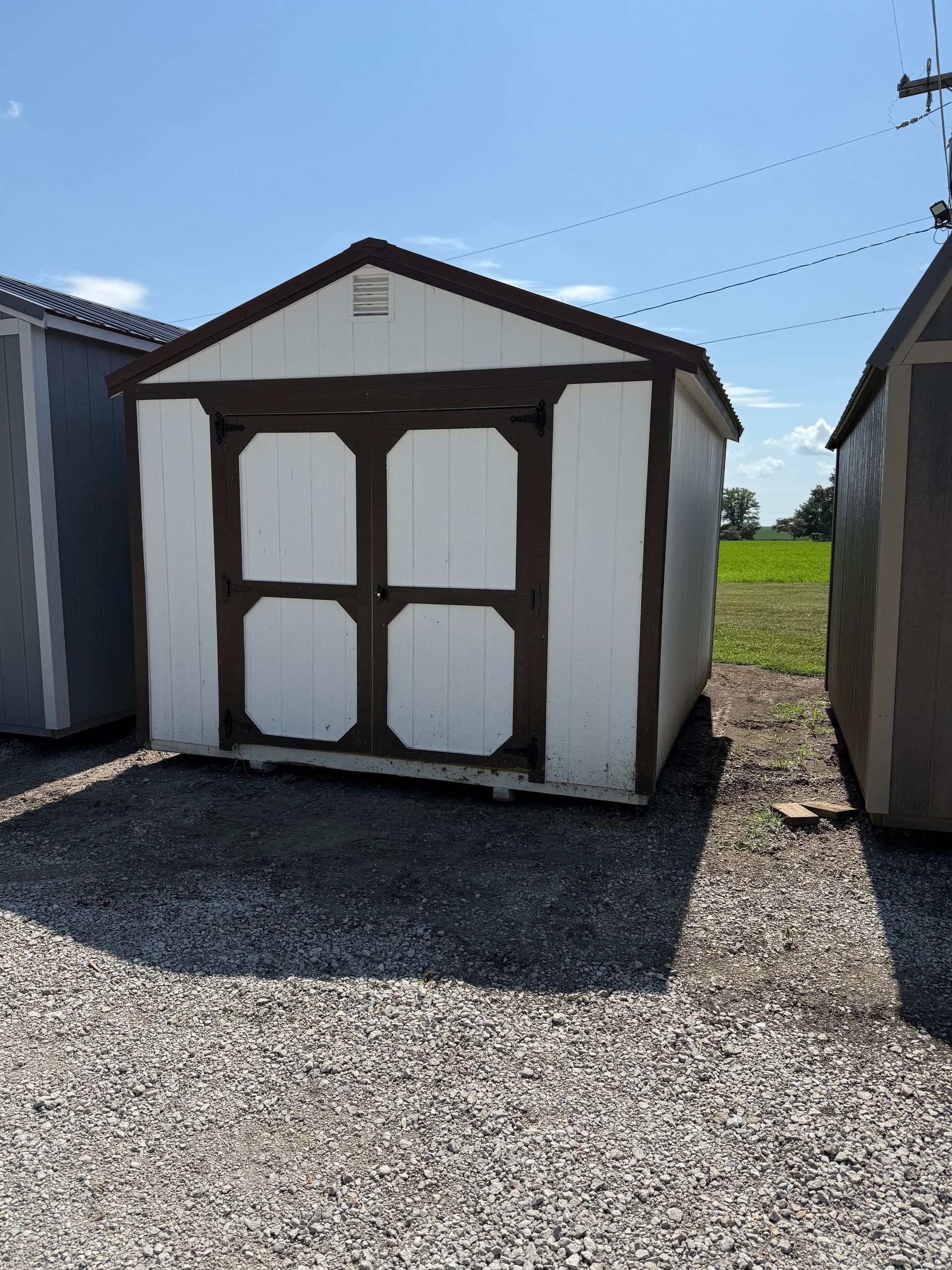 White shed with brown trim, double doors, and gravel ground.