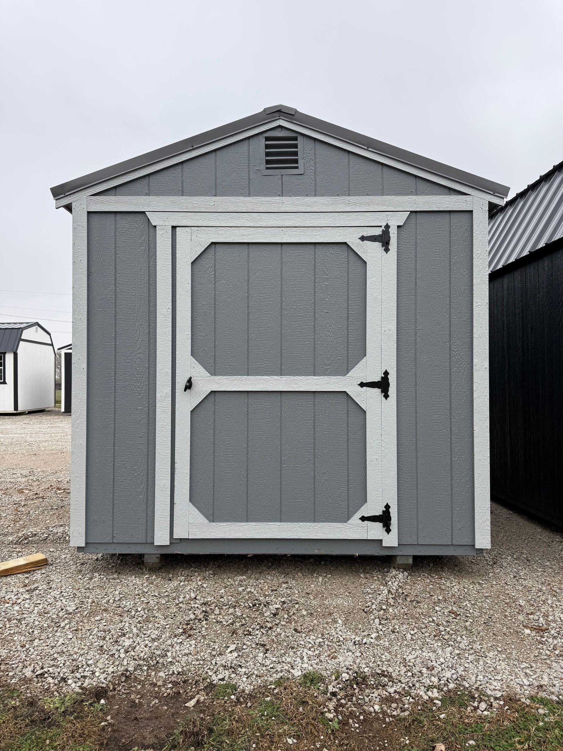 Gray shed with white trim, a single door, and a vent under the roof. It is set on gravel.