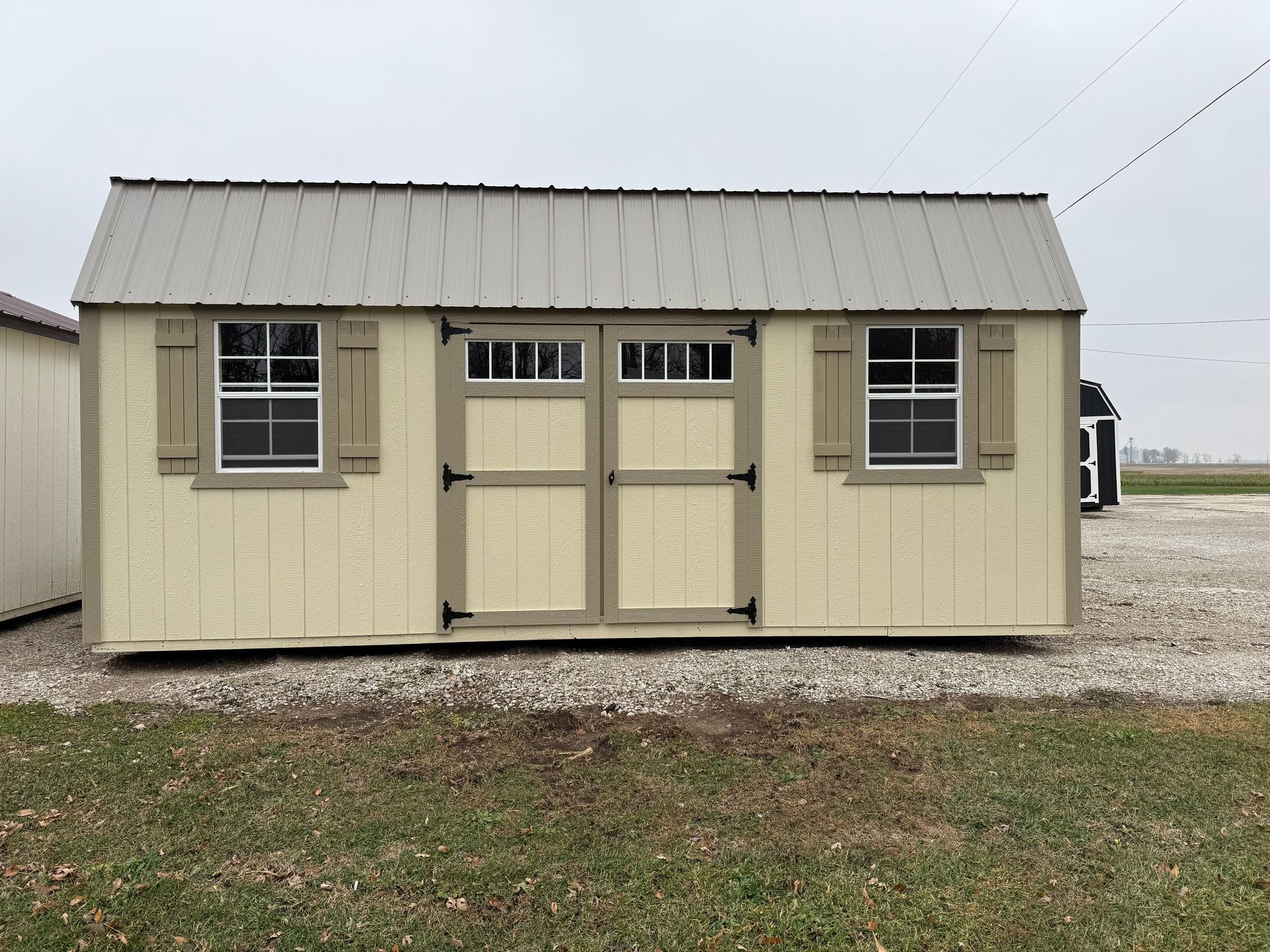 Tan shed with a metal roof, two windows with shutters, and double doors.