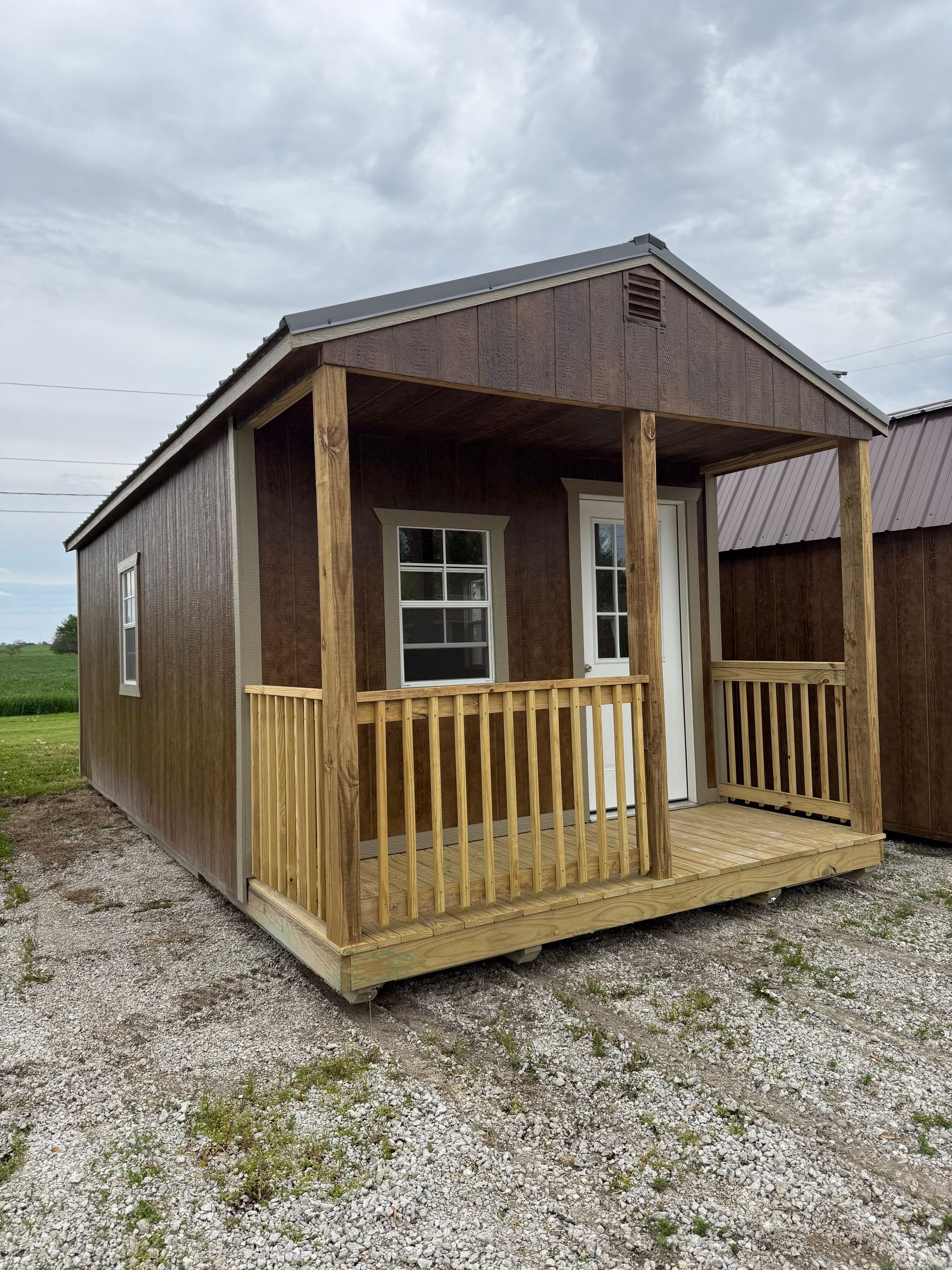 Brown shed with a porch and wooden railings sits on a gravel lot under a cloudy sky.