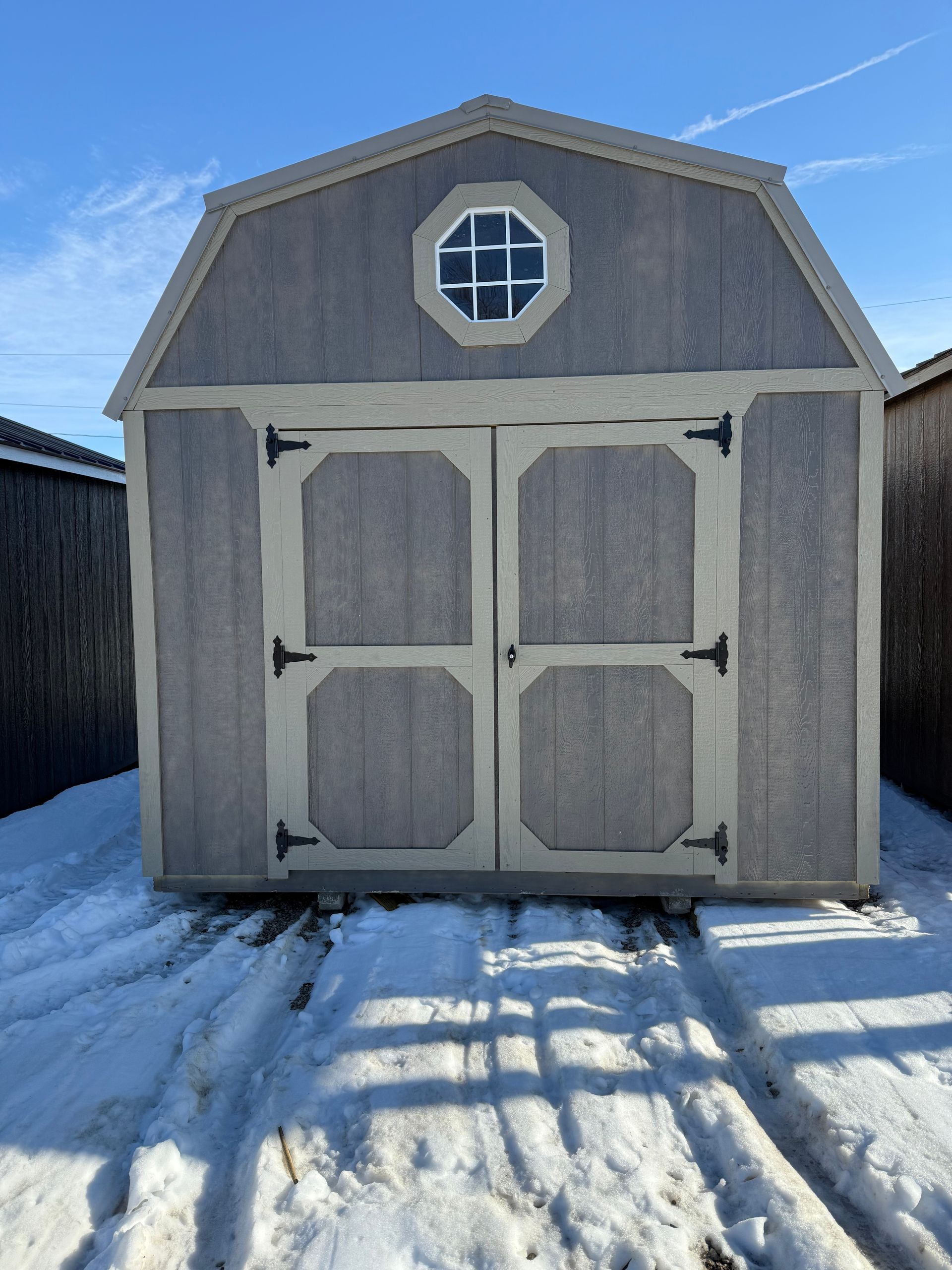 Grey barn shed with cream trim and an octagonal window, in a snowy setting.