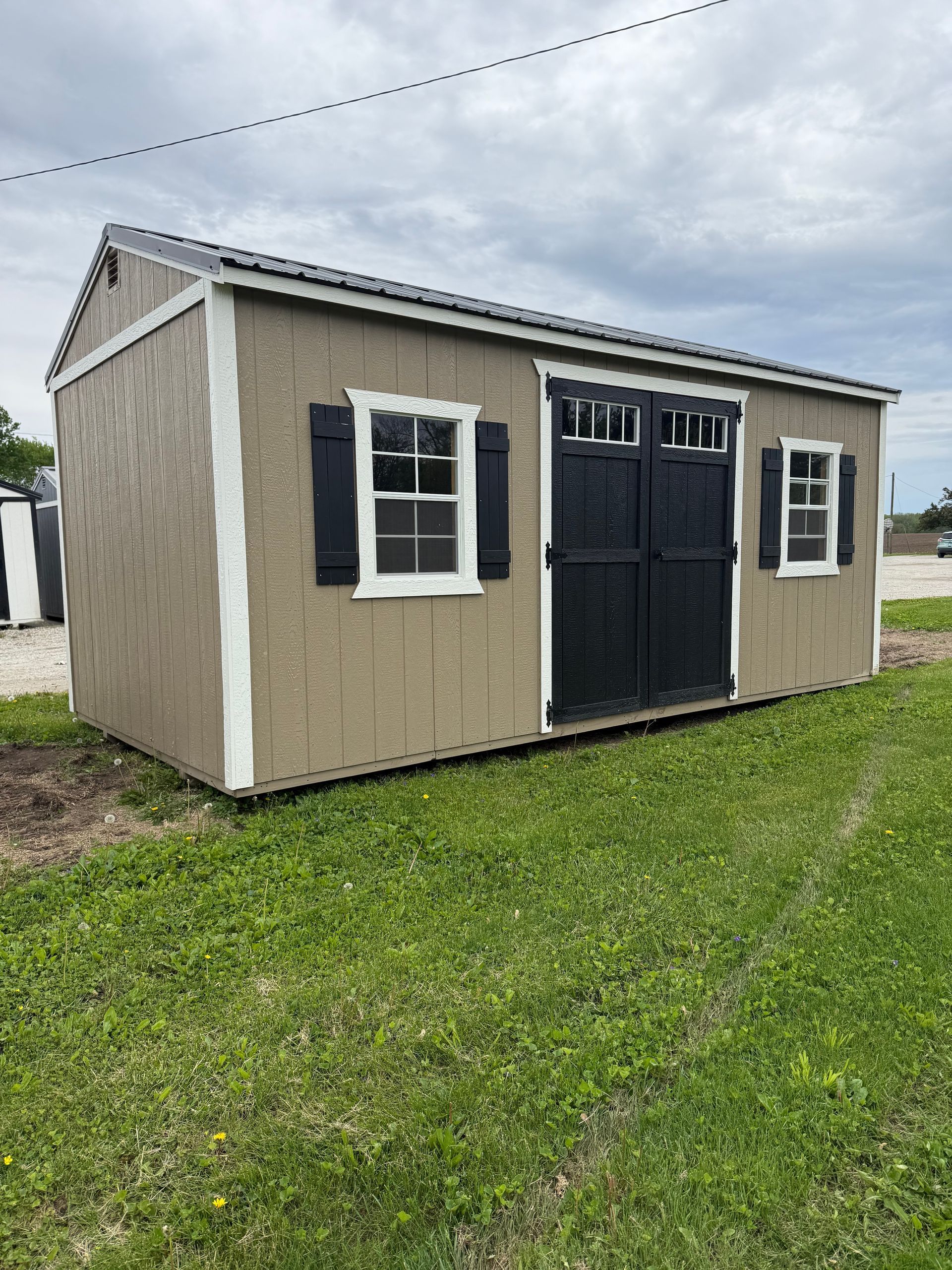 Tan shed with black shutters, white trim, and a gray roof on a green lawn.