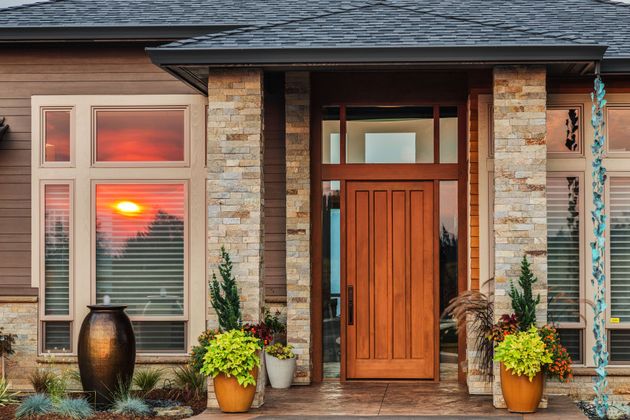 Front entrance of a modern home with a wooden door, stone pillars, potted plants, and a sunset reflection in the window