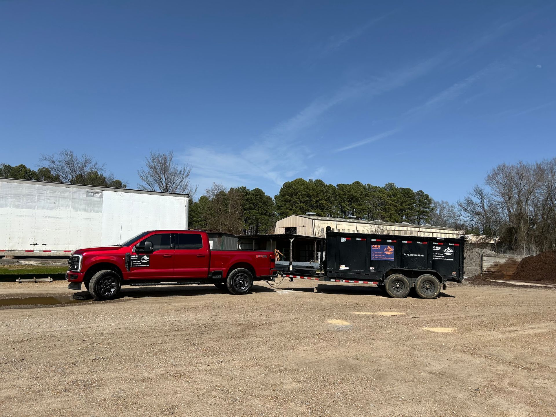 Red pickup truck parked beside a black utility trailer on a dirt lot under a clear blue sky.