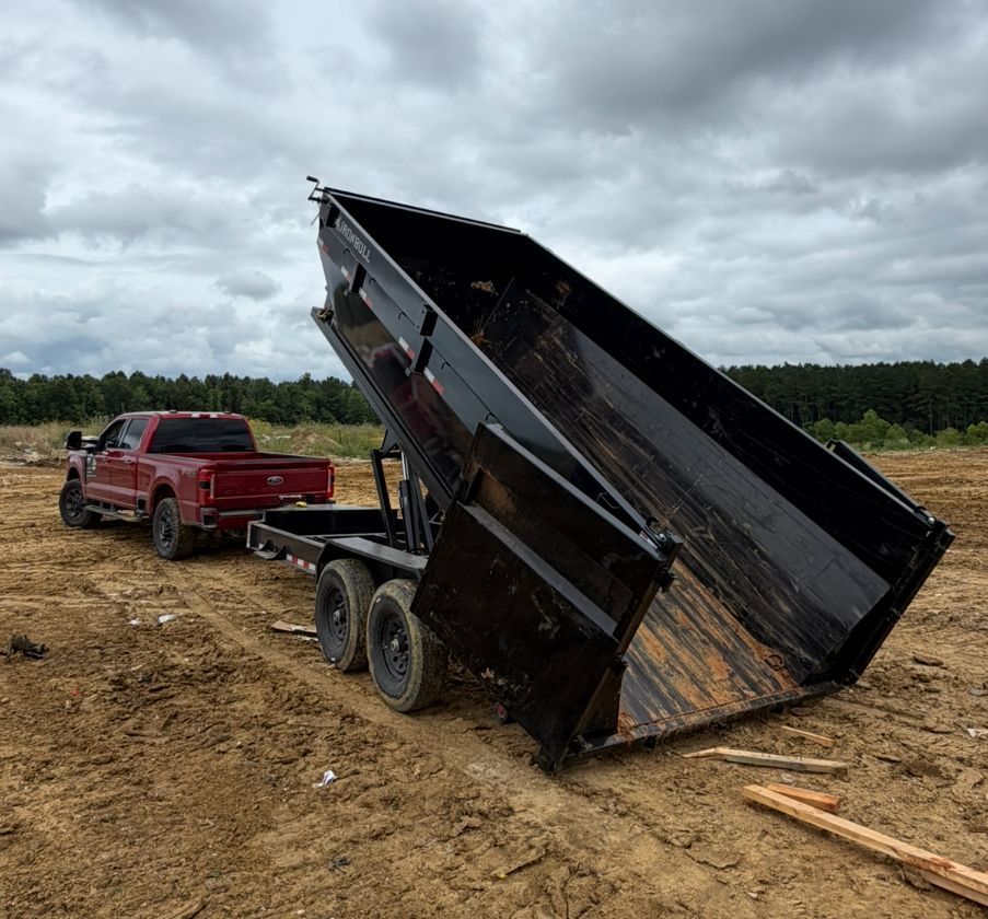 Red pickup truck towing a tilted black dump trailer in a muddy field under cloudy skies