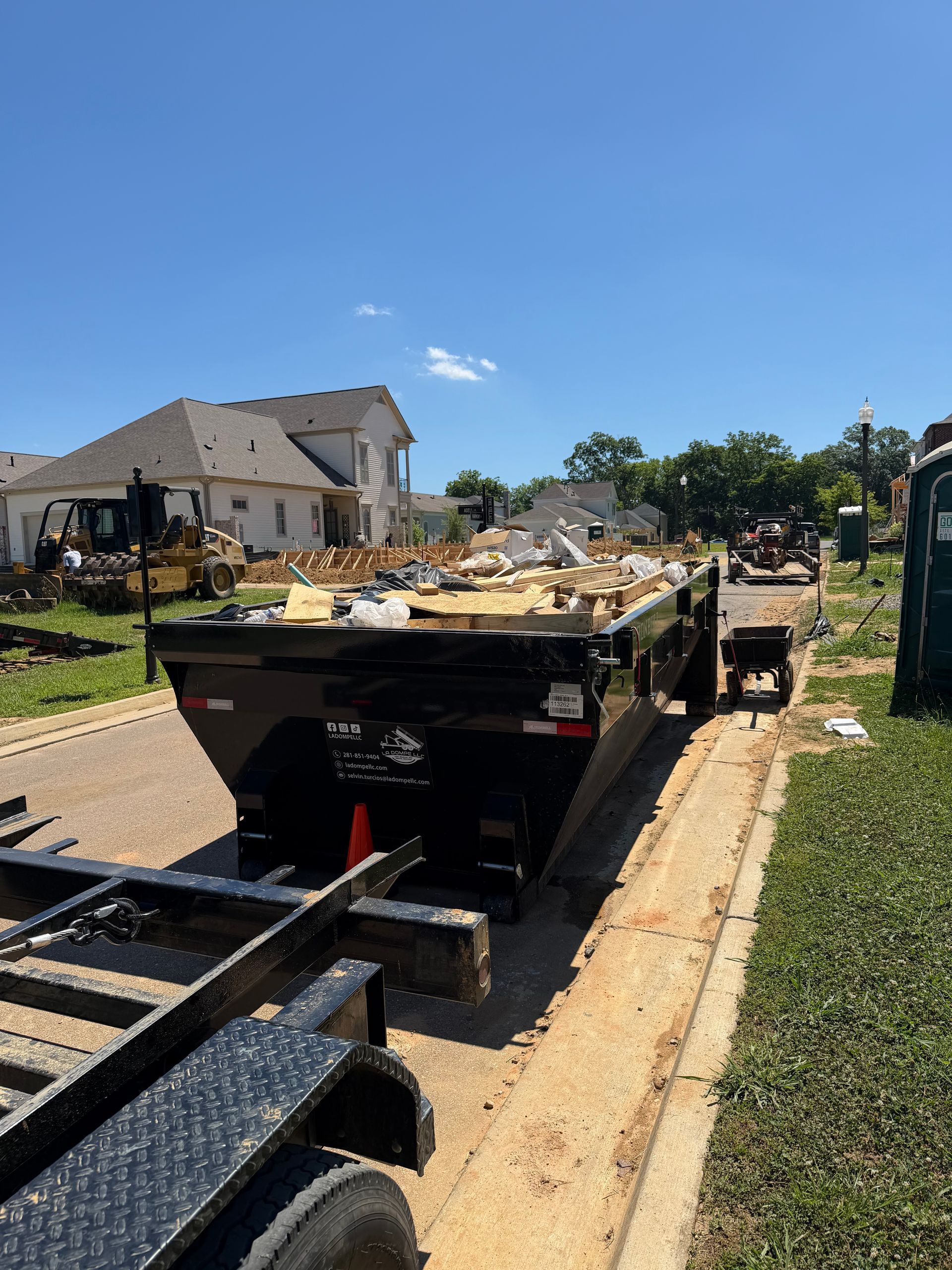 Flatbed trailer loaded with debris on a sunny residential street, with houses and utility poles nearby