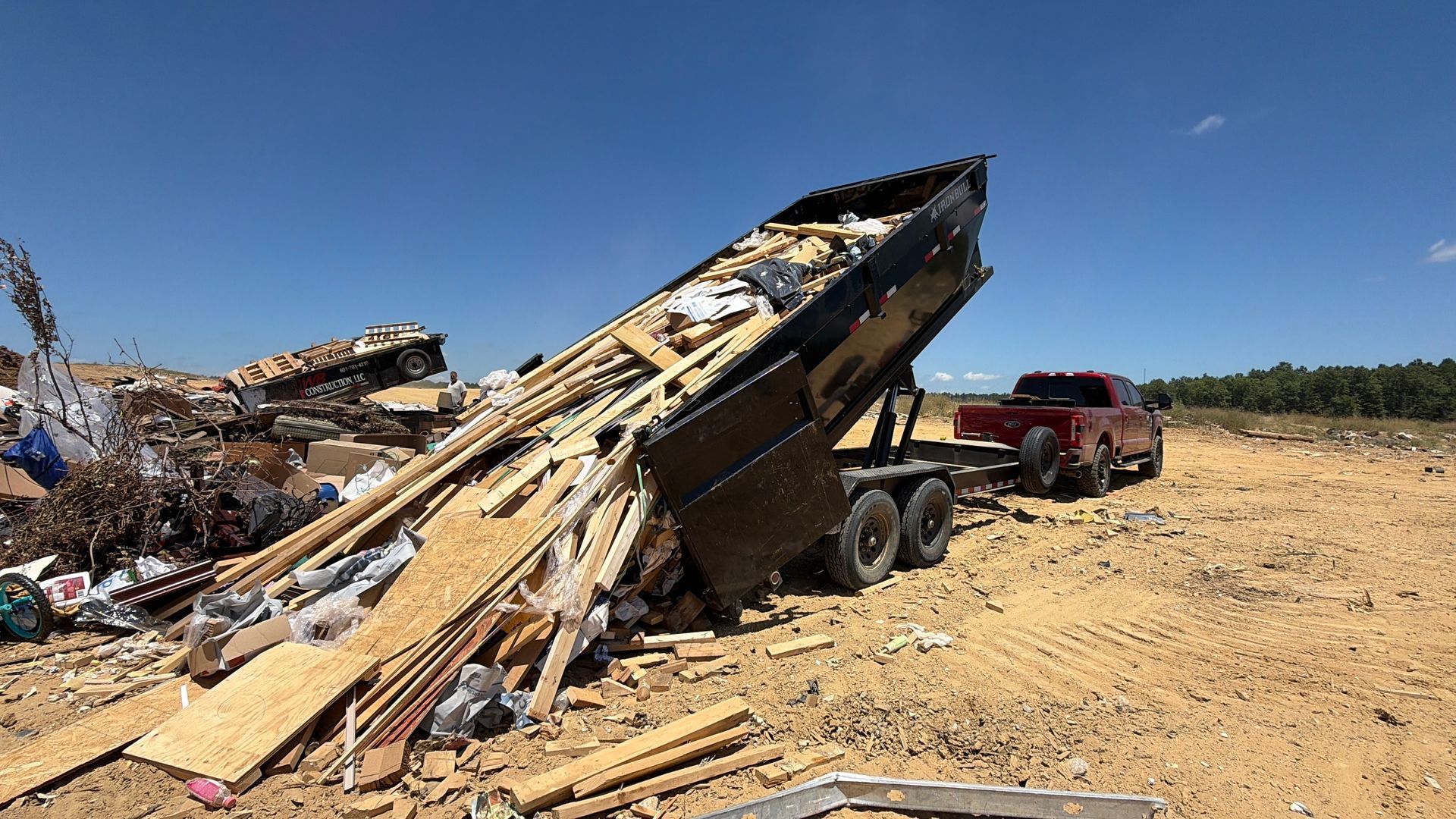 Collapsed wooden structure on a trailer in a dusty lot, with debris and a red truck nearby