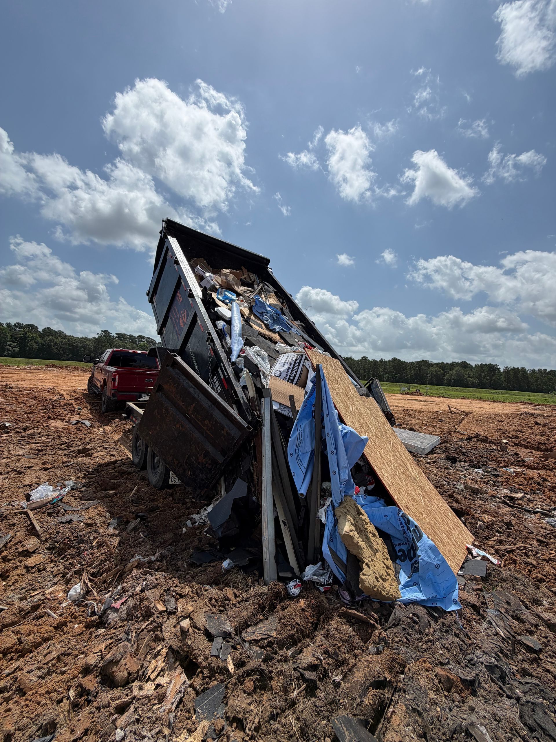 Overturned dump truck in a muddy field under a blue sky with clouds
