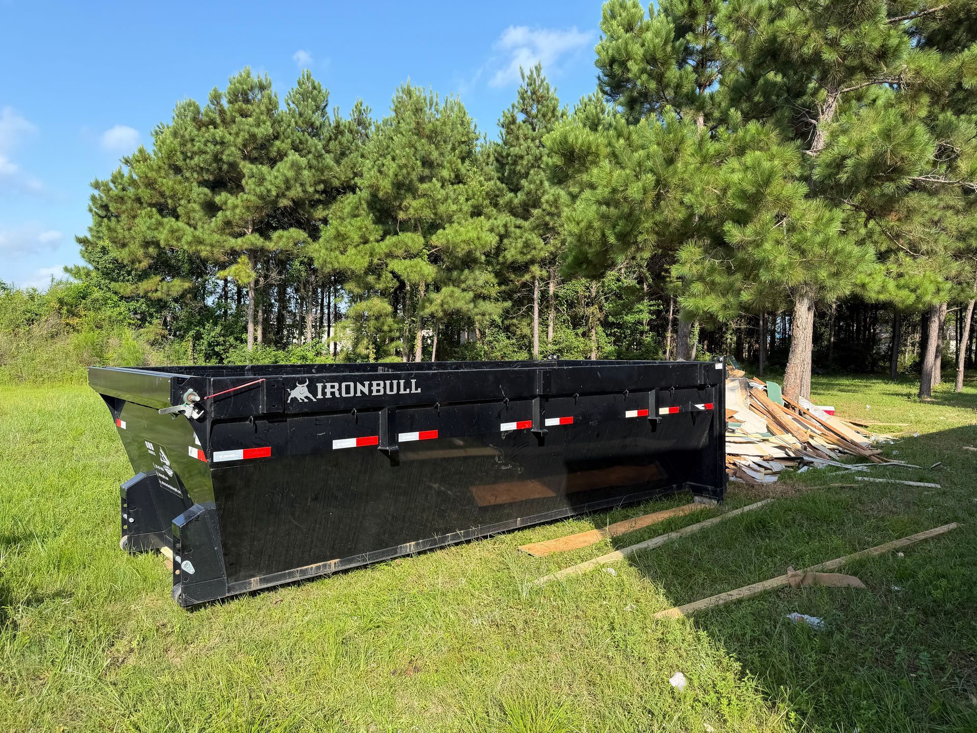 Black dump trailer in a grassy field beside trees under a blue sky