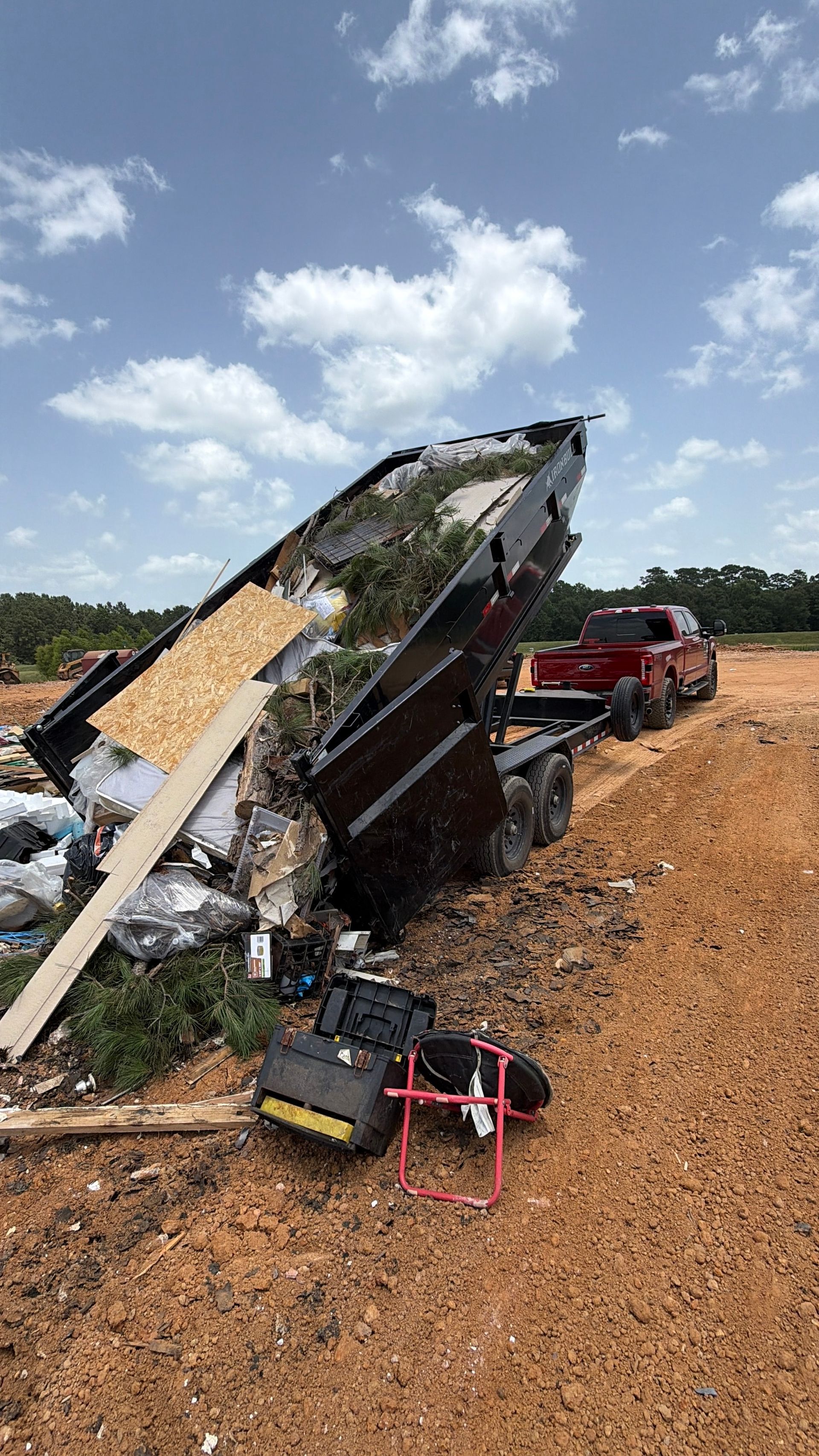 Overturned red truck hauling debris on a muddy roadside under a partly cloudy sky