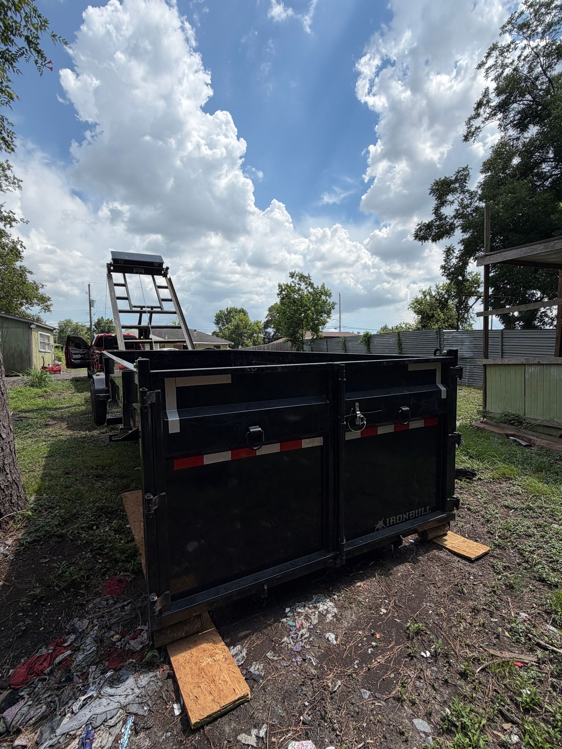 Overturned black utility trailer on gravel driveway under a cloudy sky, with a ladder nearby and debris around it