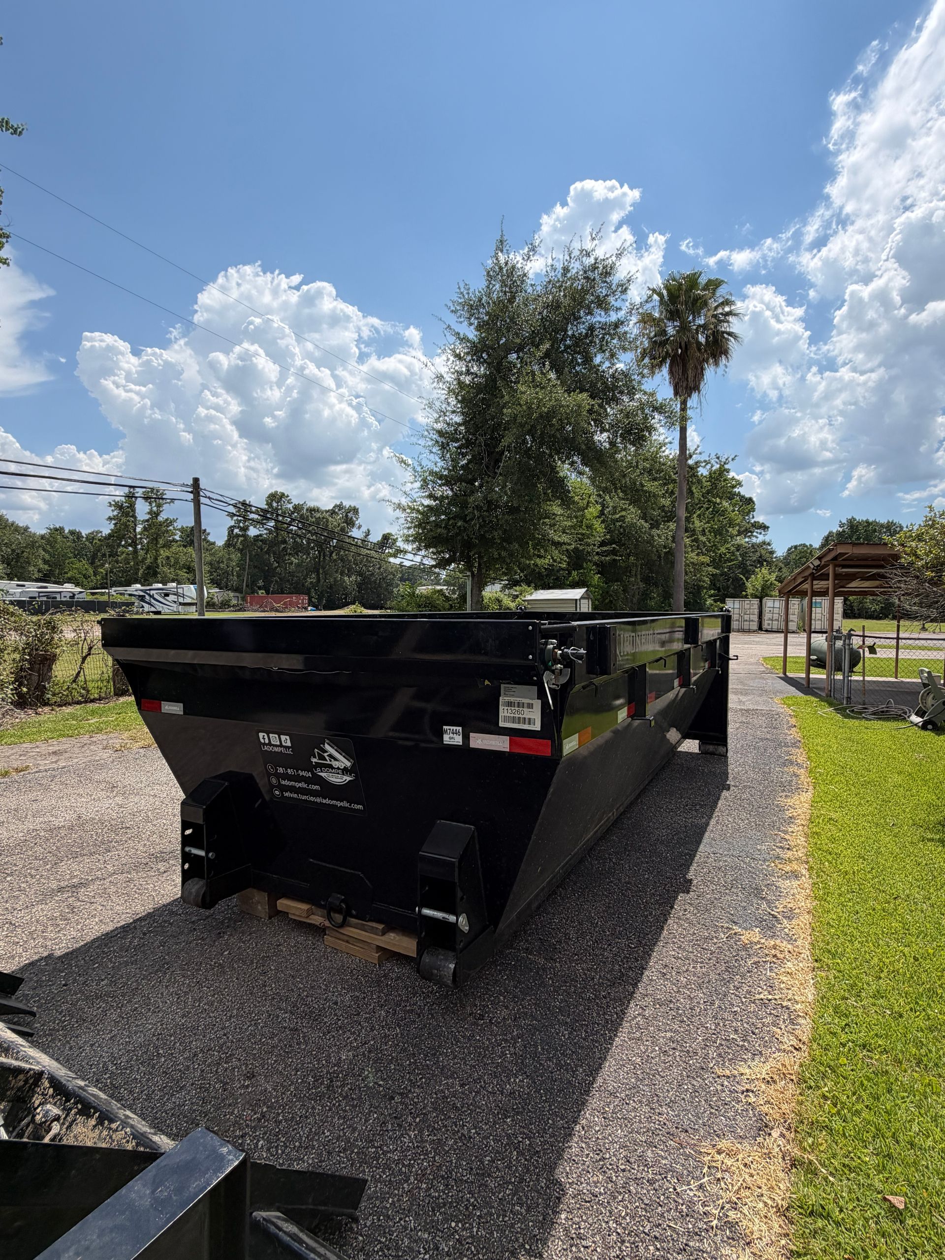 Black metal dumpster on gravel driveway under a bright blue sky with clouds and trees in the background