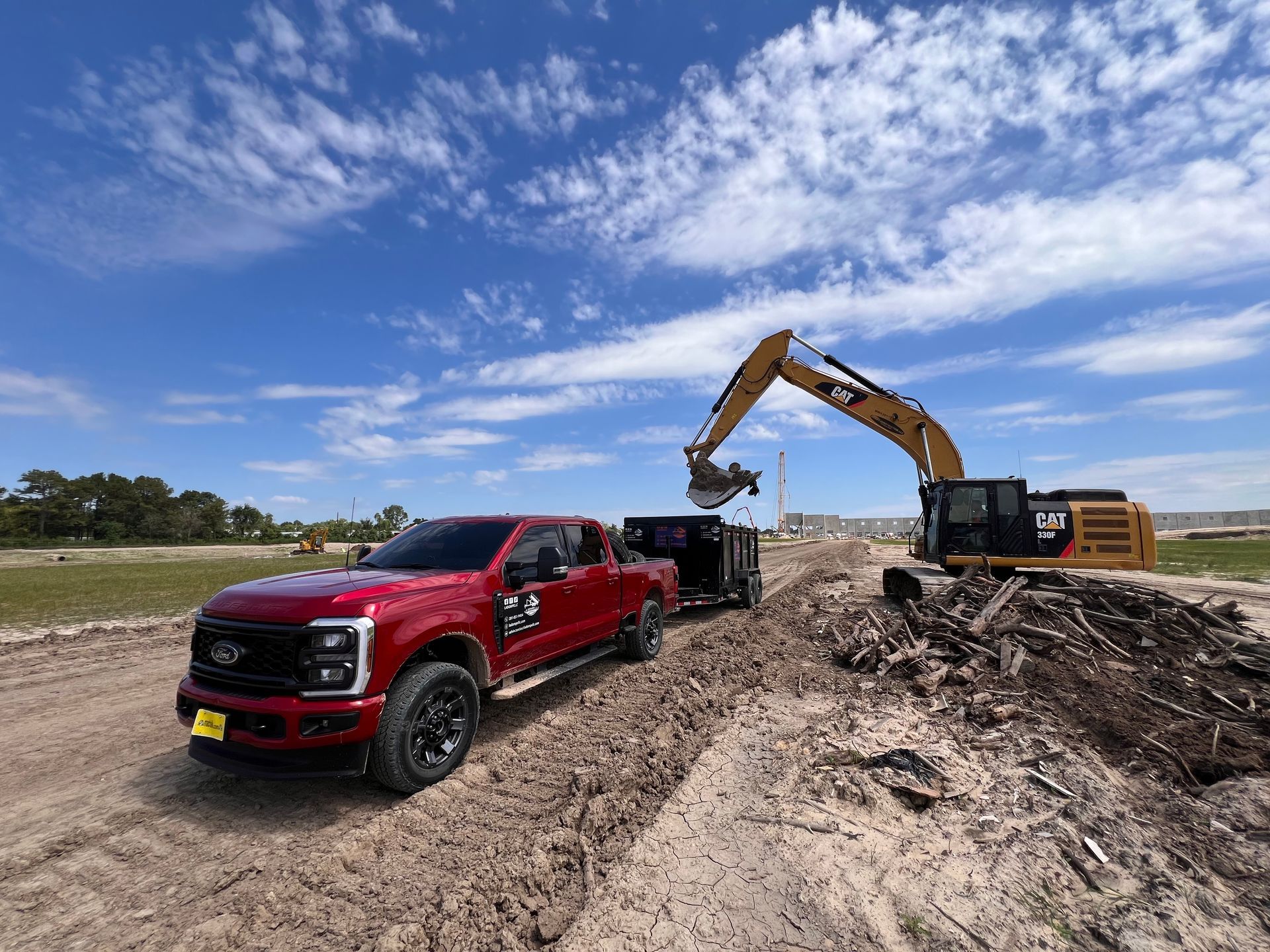 Red pickup truck beside excavator at a dirt construction site under a cloudy blue sky