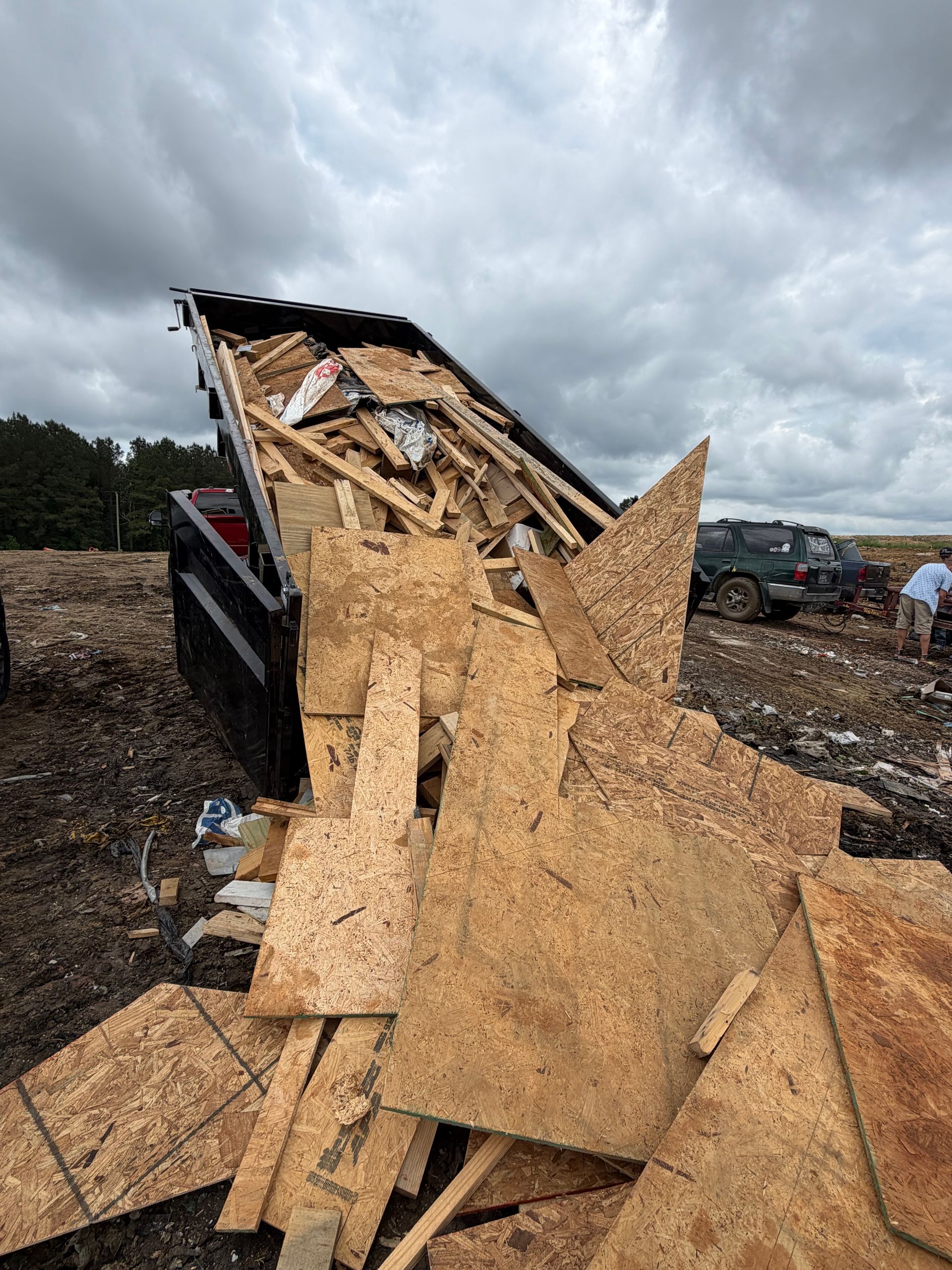 Damaged black trailer dumping broken plywood and debris in a muddy lot under cloudy skies