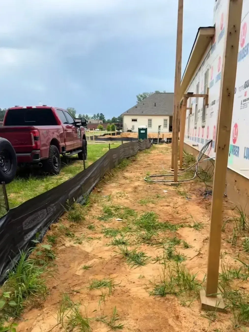 Construction site beside a house with wooden posts, dirt ground, and a red pickup truck parked nearby