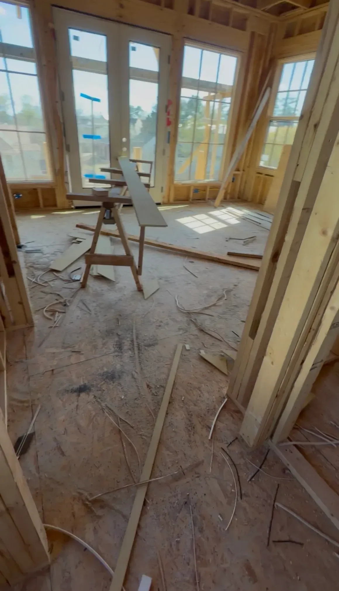 Interior of a house under construction with exposed framing, debris, and a ladder near large windows.
