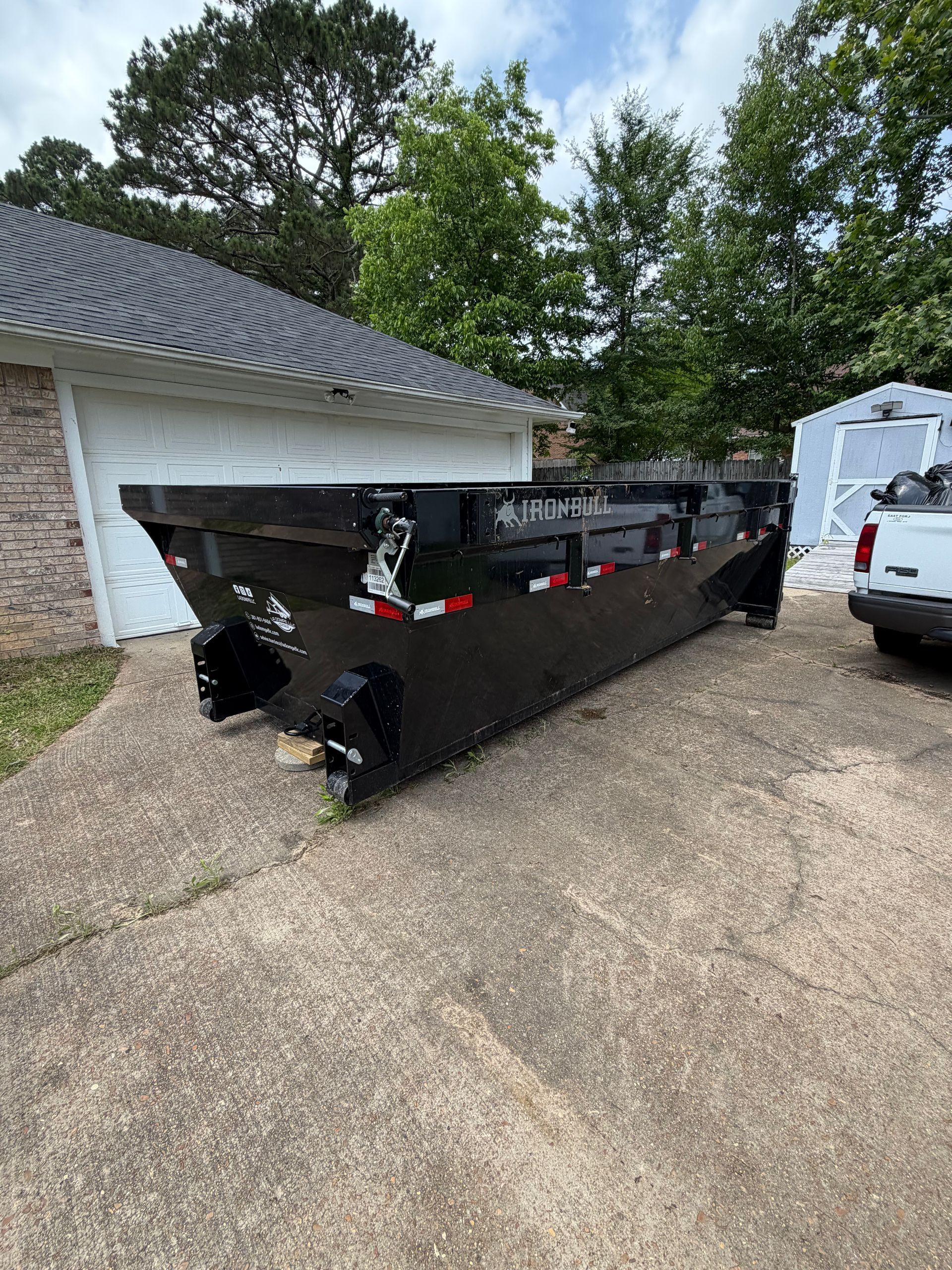 Black dumpster filled with debris beside a garage and white truck on a driveway.