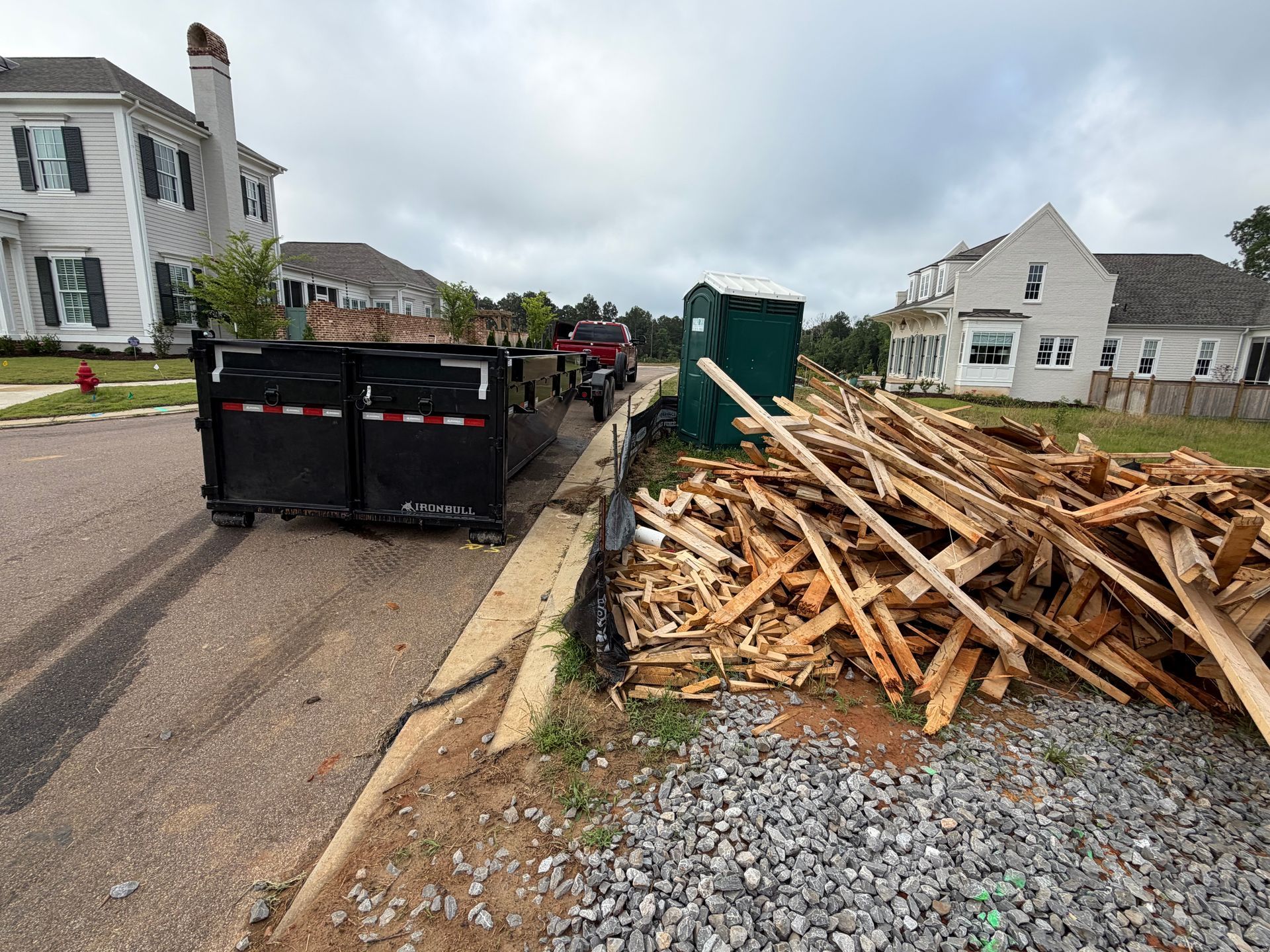 Large pile of broken wood beside a road with a black dumpster and houses in the background