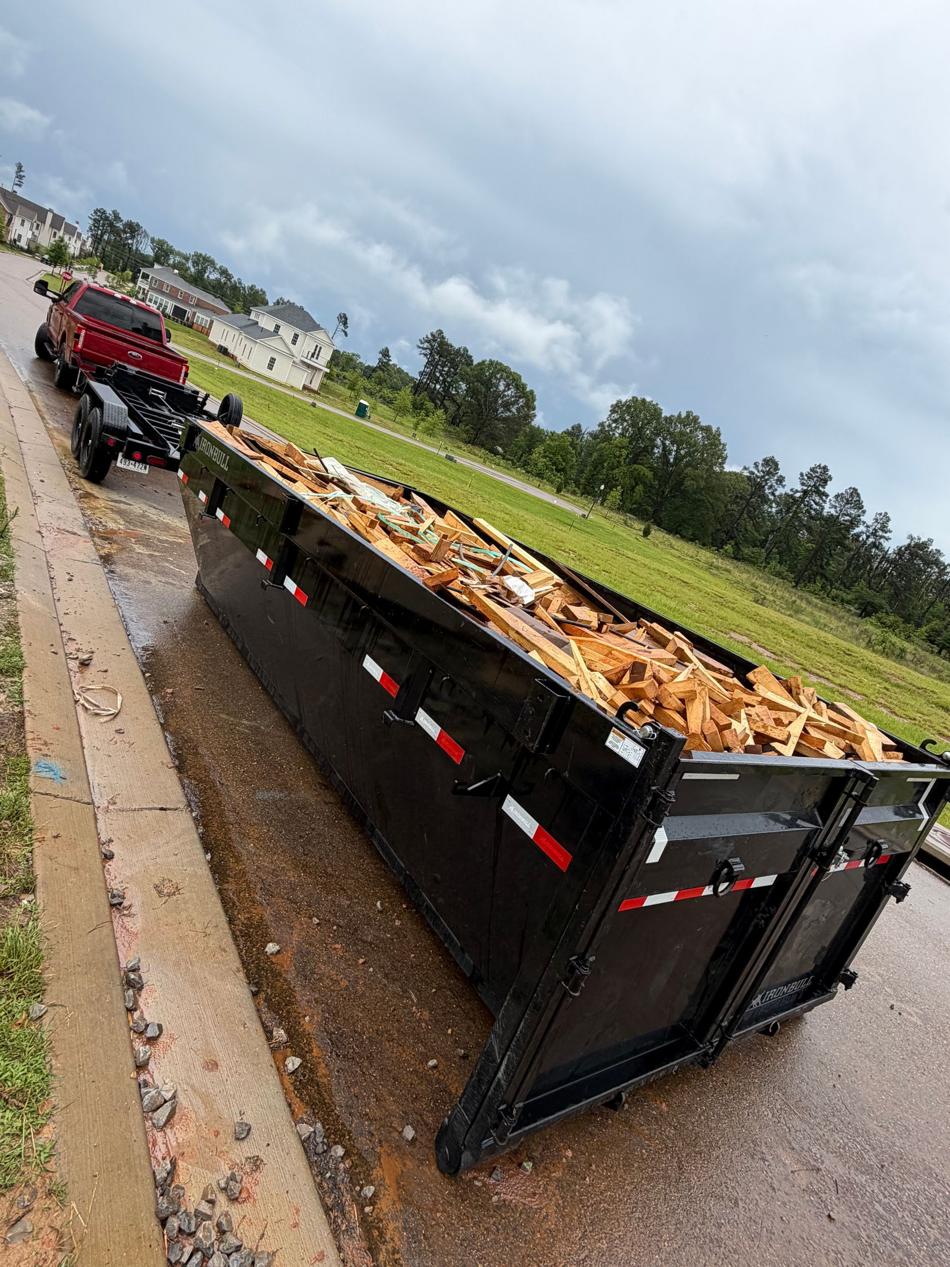 Reddish-brown dumpster loaded with debris on a wet roadside, with a red truck behind it and fields in the background