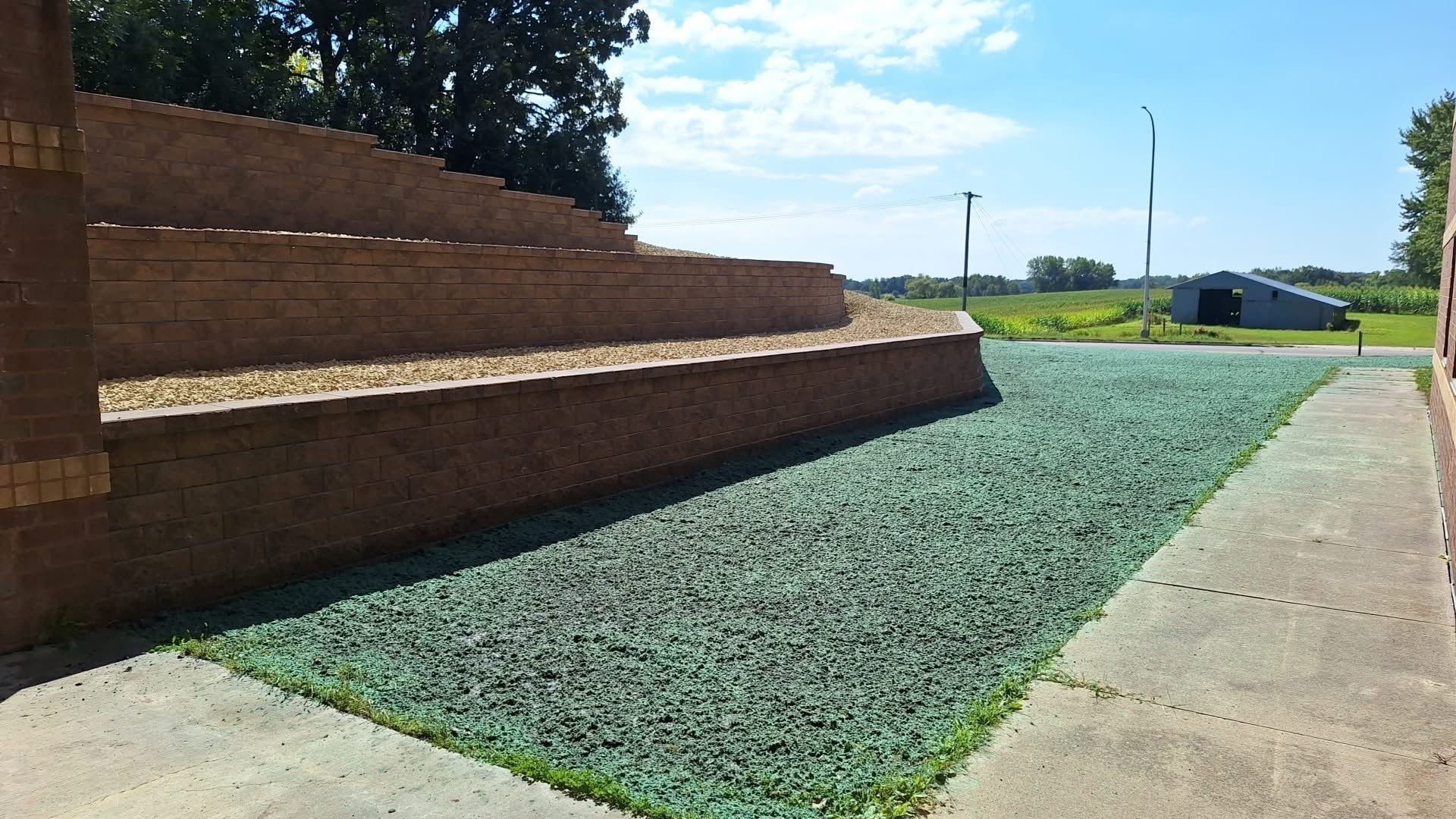Low brick tiered wall with green ground cover next to a concrete walkway, rural setting with clear sky.