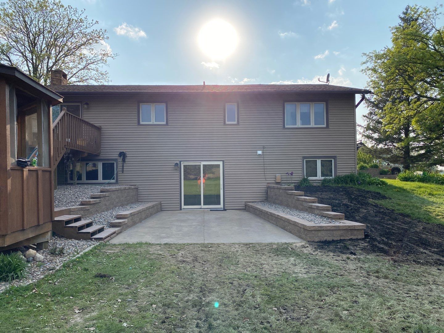 Back of a house with sliding glass door, patio, retaining walls, and sun in the sky.