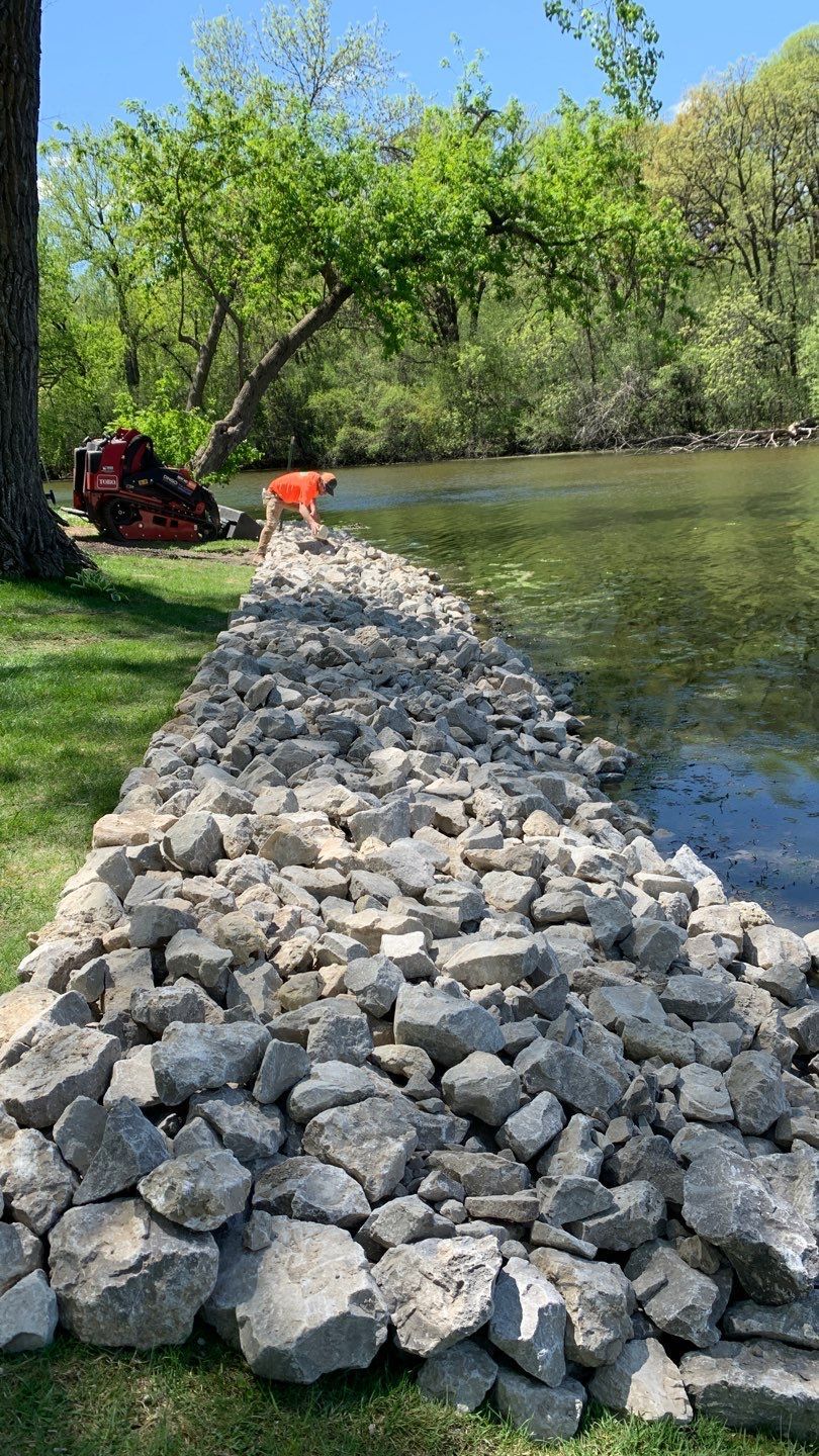 Rocks lining a lake's edge; a tree with green leaves is in the background. A small orange object is on the grass.