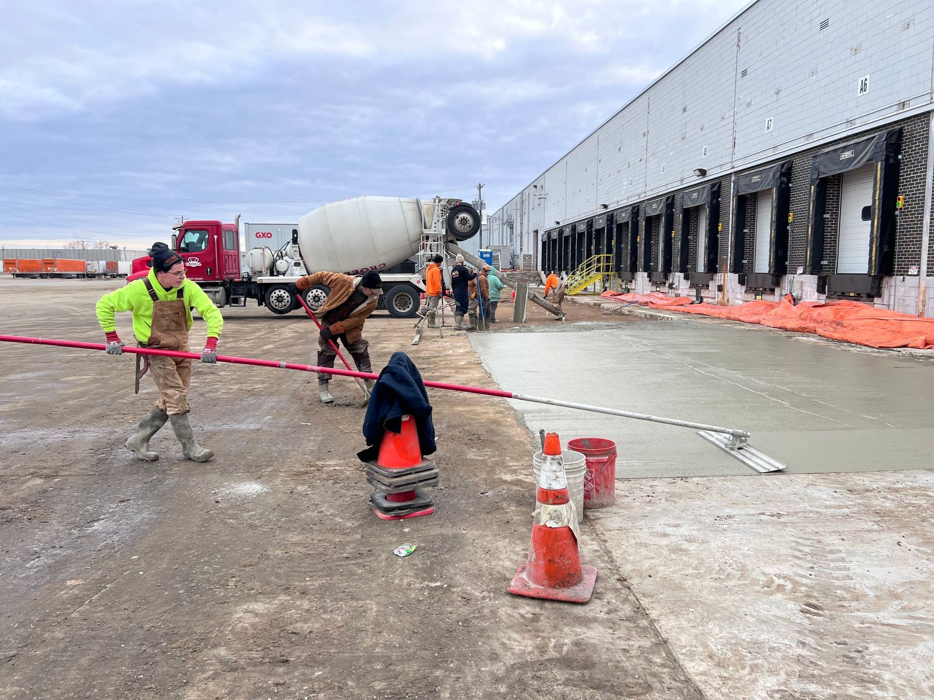 Construction workers pouring concrete at a loading dock; red cement truck, orange cones, overcast sky.