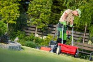 Gardener on a sloped lawn operating a red spreader, mower nearby; lush greenery in the background.