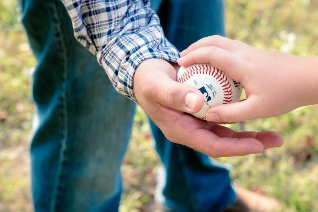 Person holding a baseball, another hand reaching to take it. Outdoors.