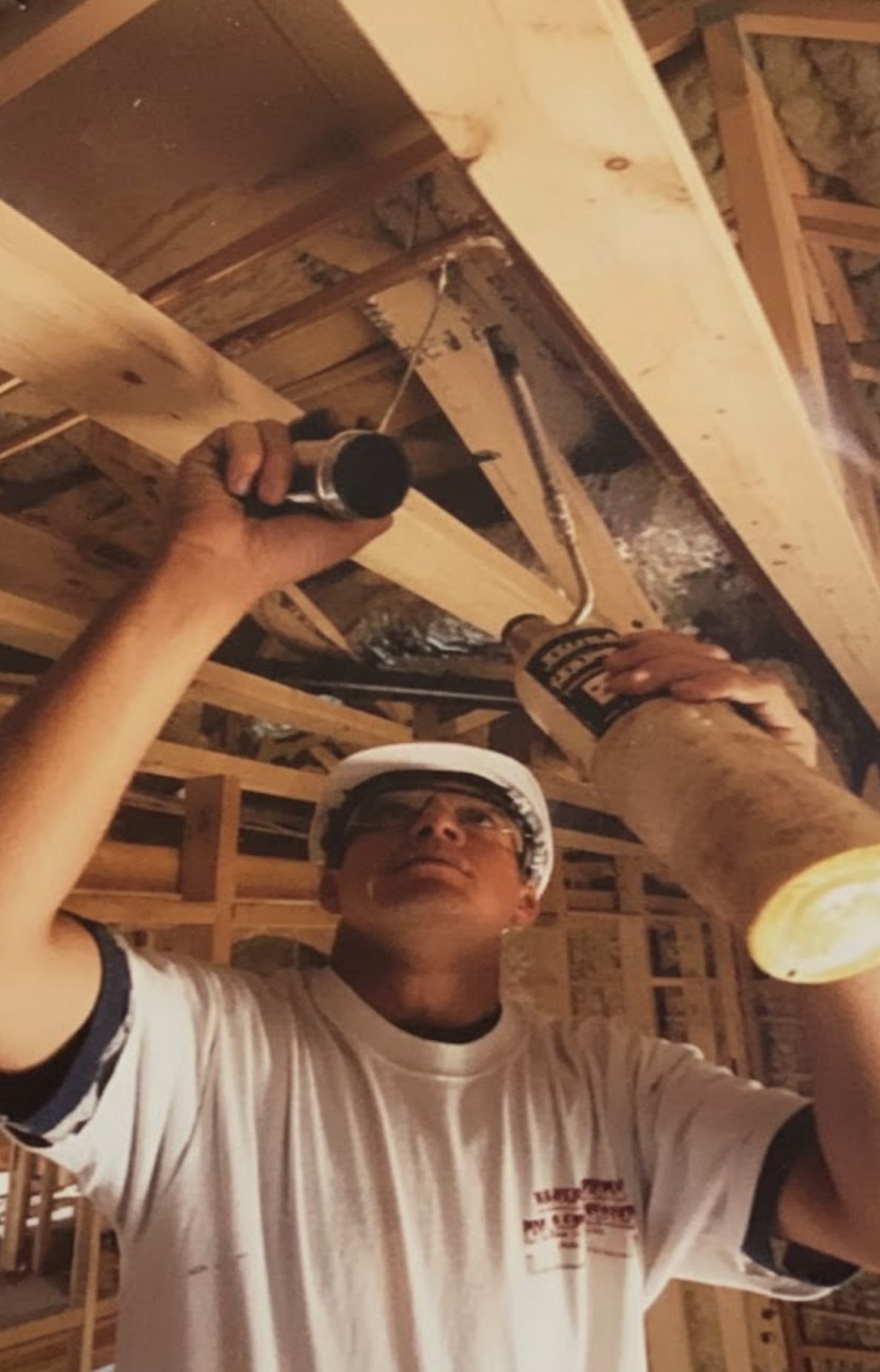 Worker installing ceiling wiring in an unfinished house, wearing a hard hat and holding tools.