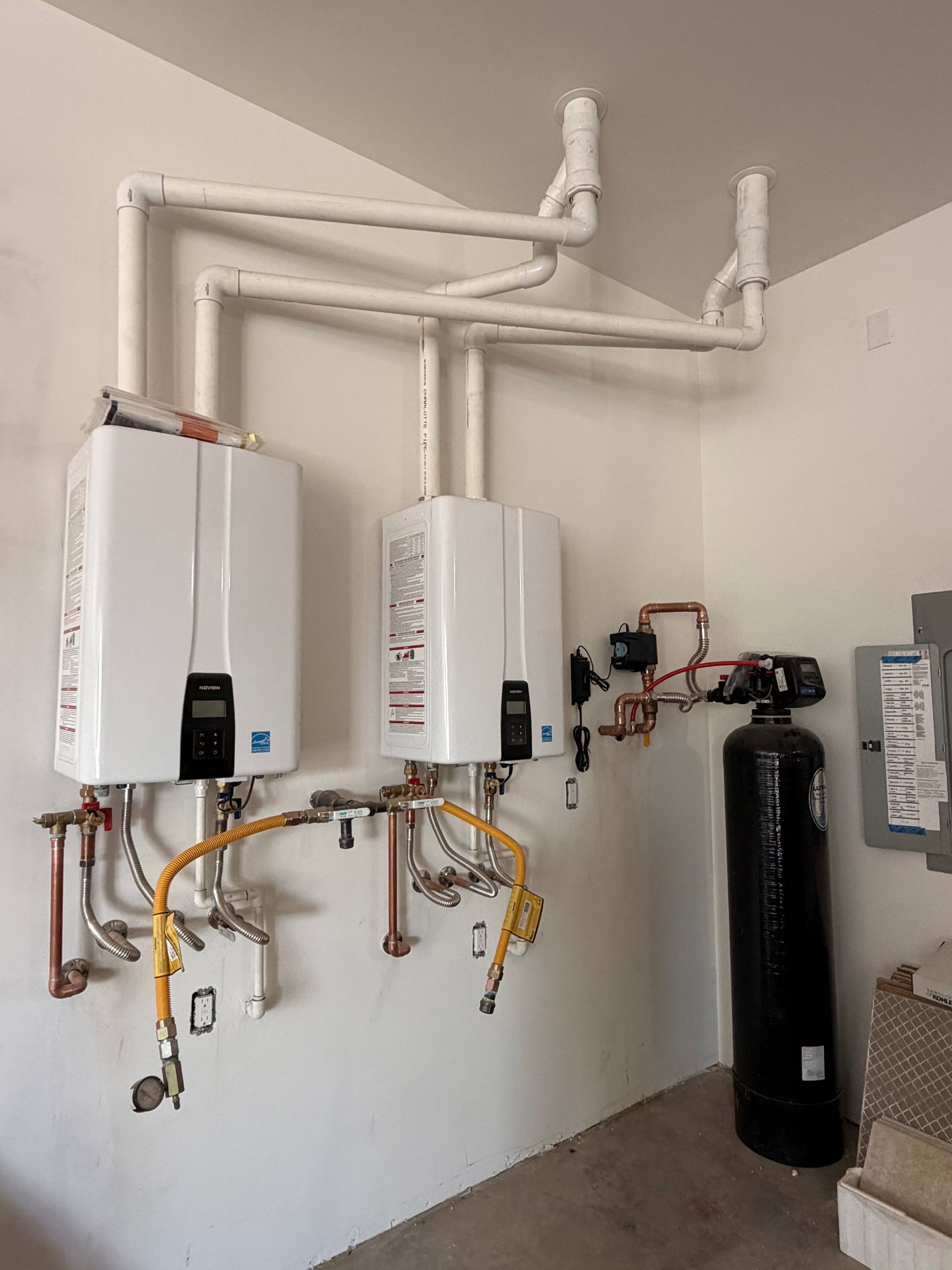 Two white wall-mounted water heaters with copper piping and a black water softener in a utility room
