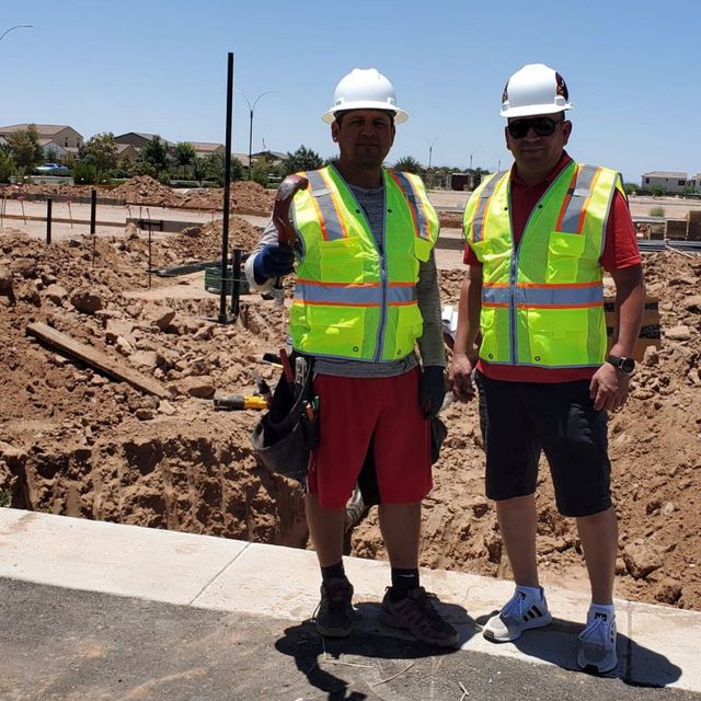 Two construction workers in hard hats and safety vests stand by a road at a dusty building site.