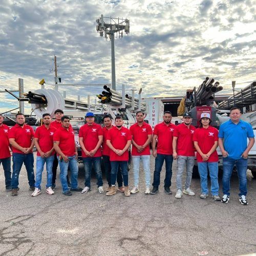 Group of workers in red shirts and one in blue posing outdoors in an industrial yard under a cloudy sky