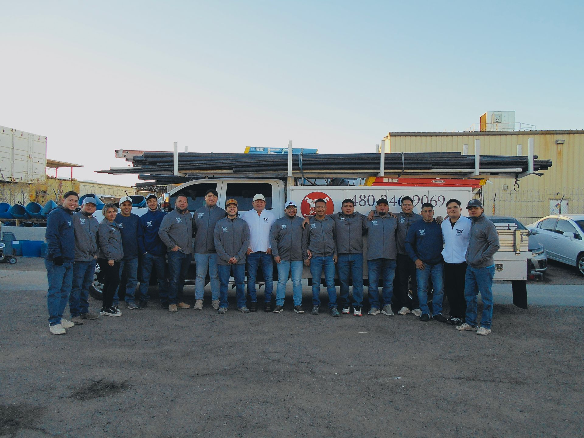 Group of workers standing together outdoors in front of industrial equipment and trucks at sunset.