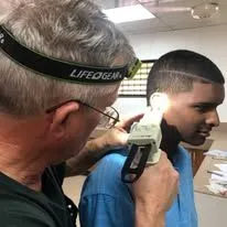 A person wearing a headlamp is examining another person's ear with a handheld device, likely related to hearing aids.
