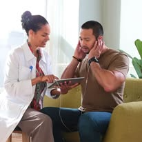 A healthcare professional shows information on a tablet to a person on a green couch, discussing Tinnitus Management Services