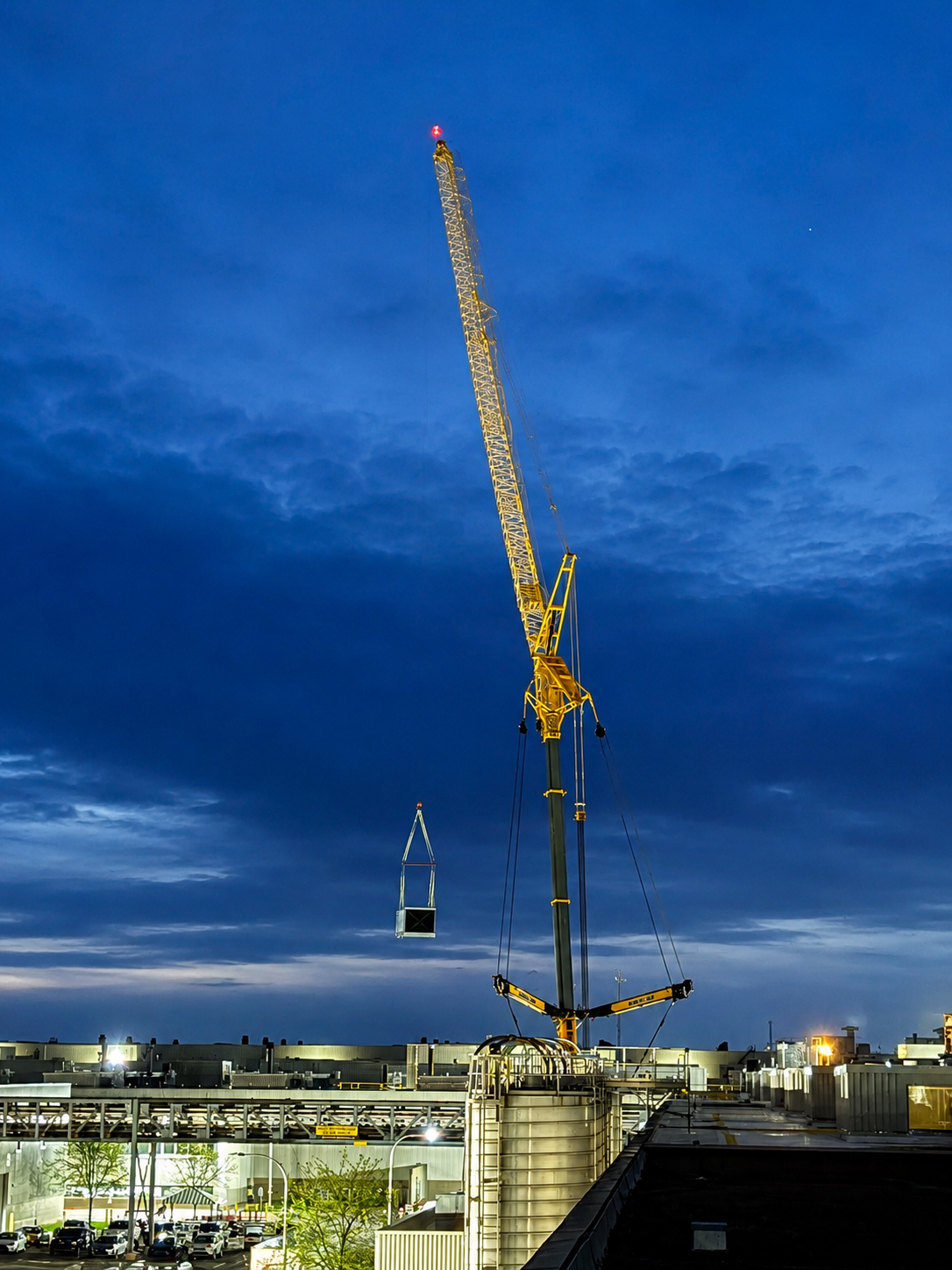 Tall construction crane at dusk hoists a hanging load above a lit building site under a blue sky.