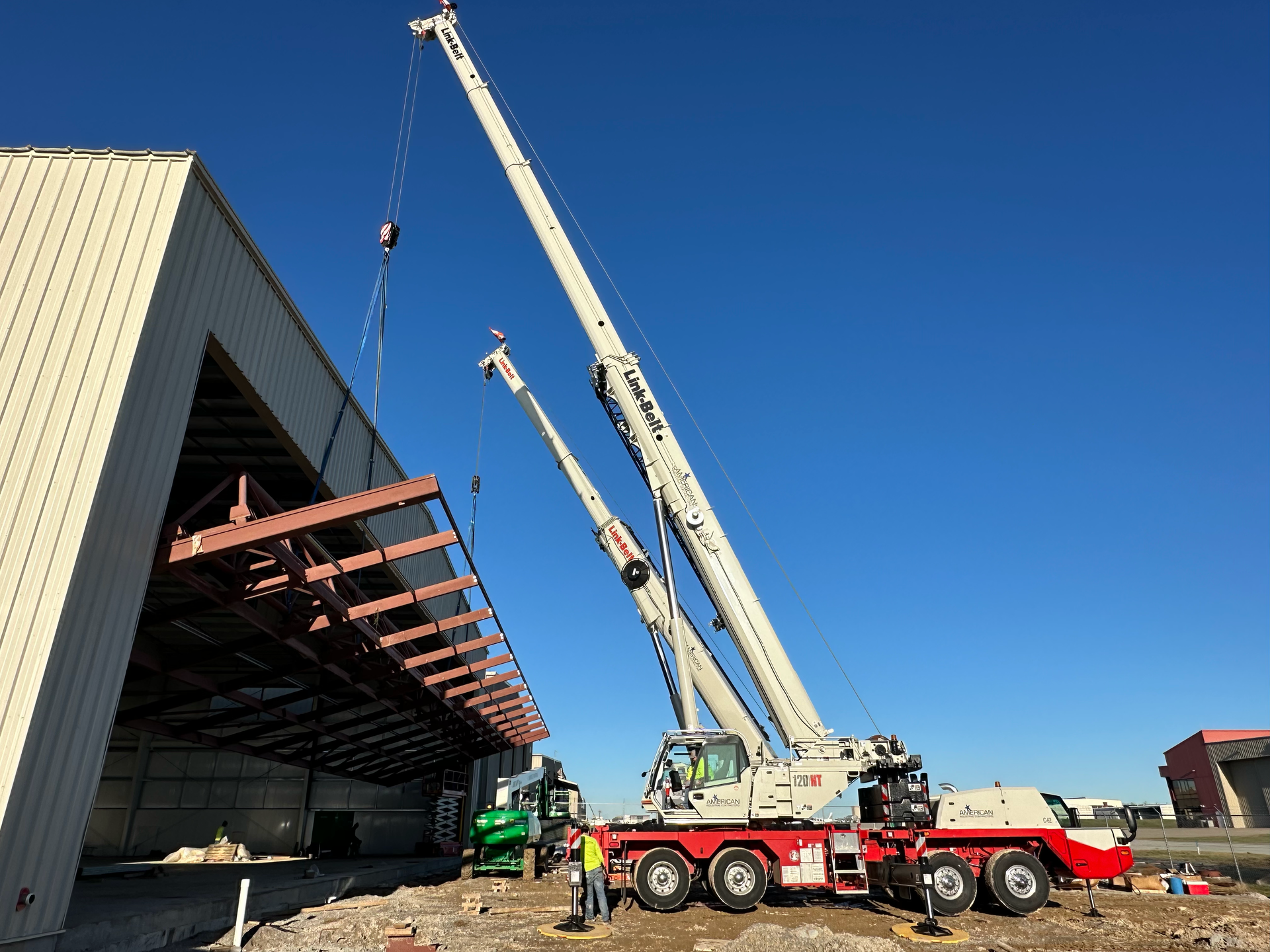 Crane trucks lifting steel beams beside an industrial warehouse under a clear blue sky