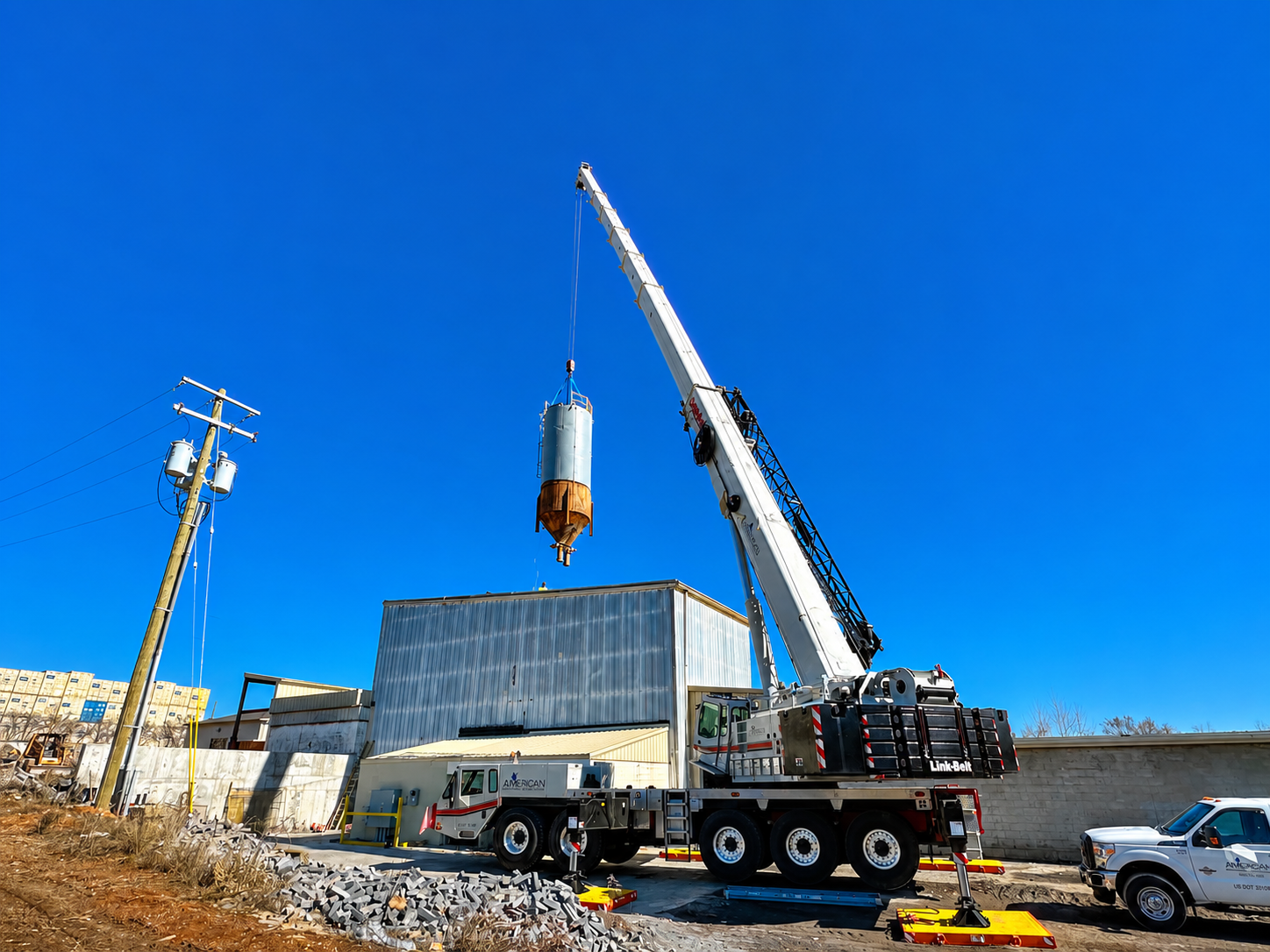 Crane lifting a large cylindrical tank beside a truck at an industrial site under a bright blue sky
