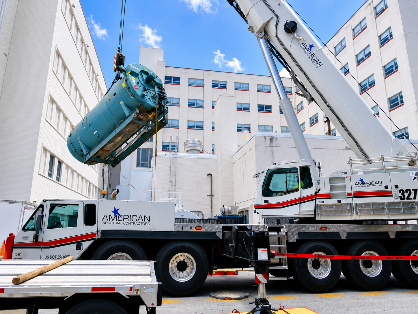 Crane lifting a blue machine between white buildings at an industrial site