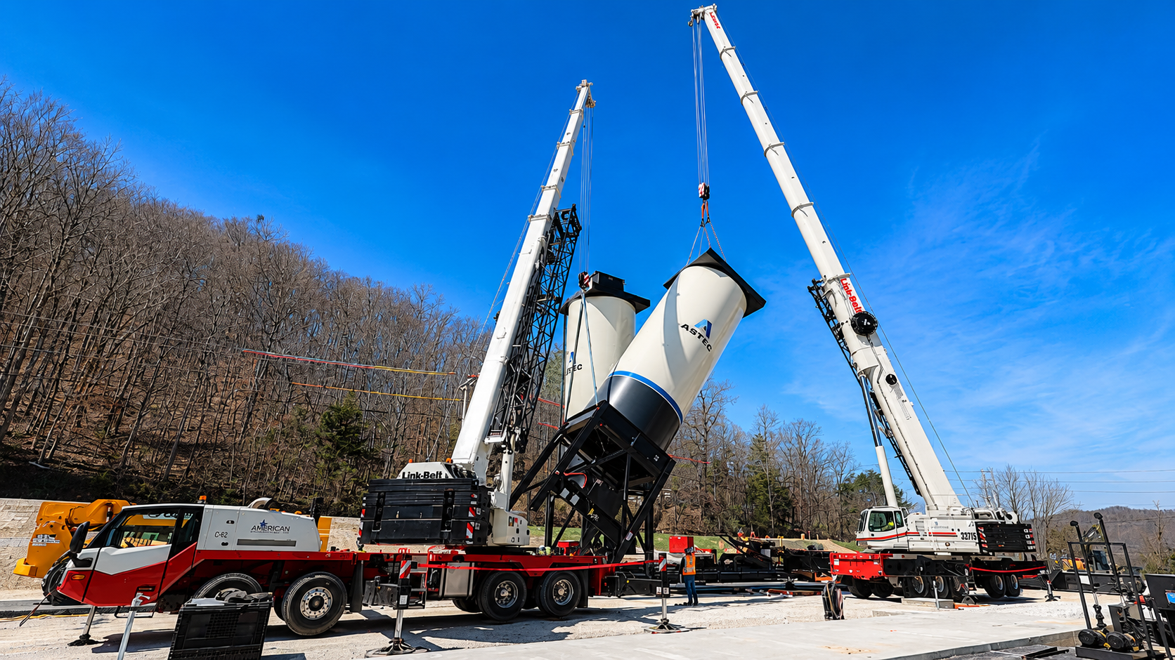 Two cranes lifting a large silver tank onto a flatbed truck at a construction site under a blue sky