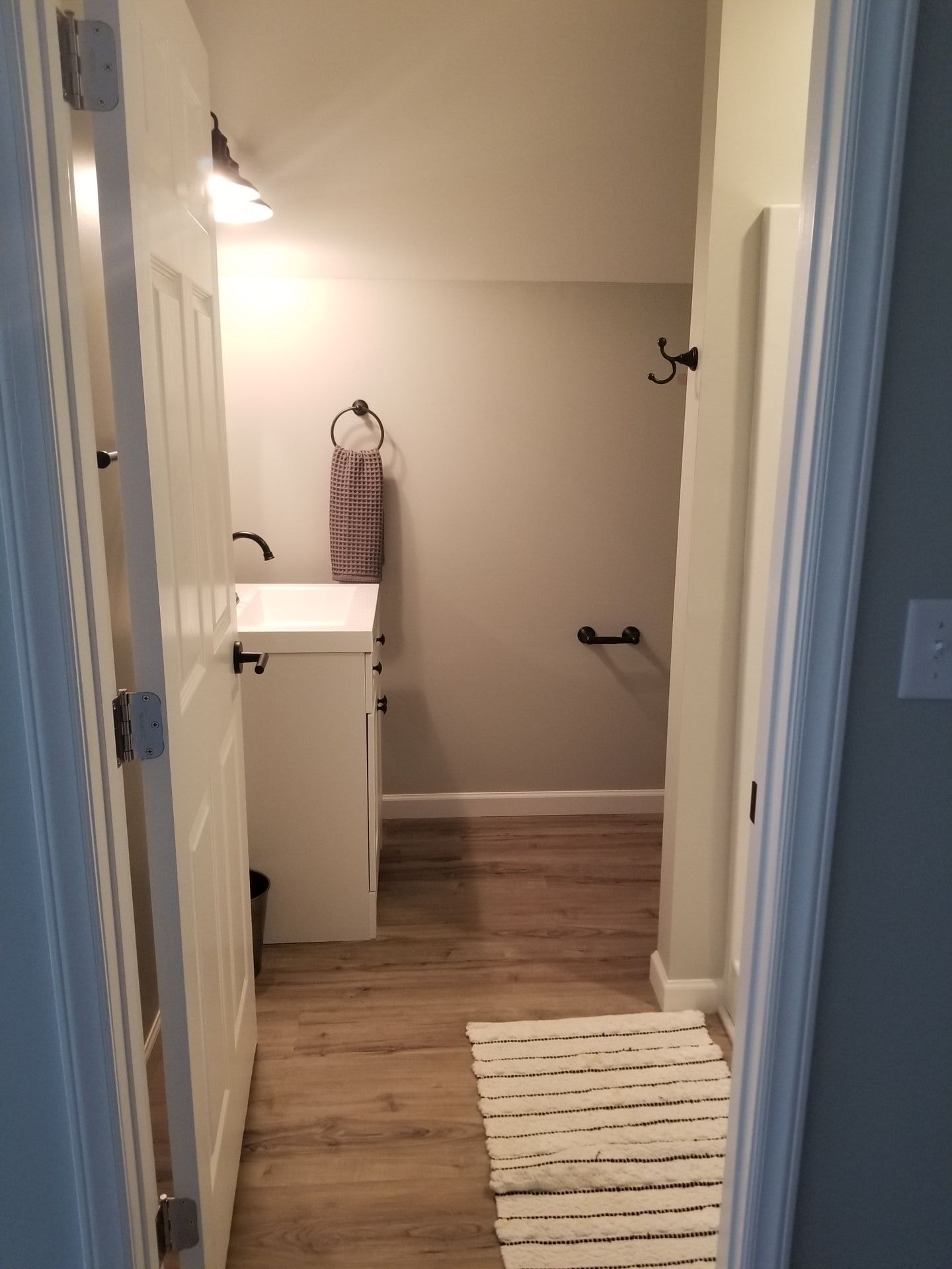 Narrow bathroom interior with white door and vanity, wood-look floor, and gray walls.