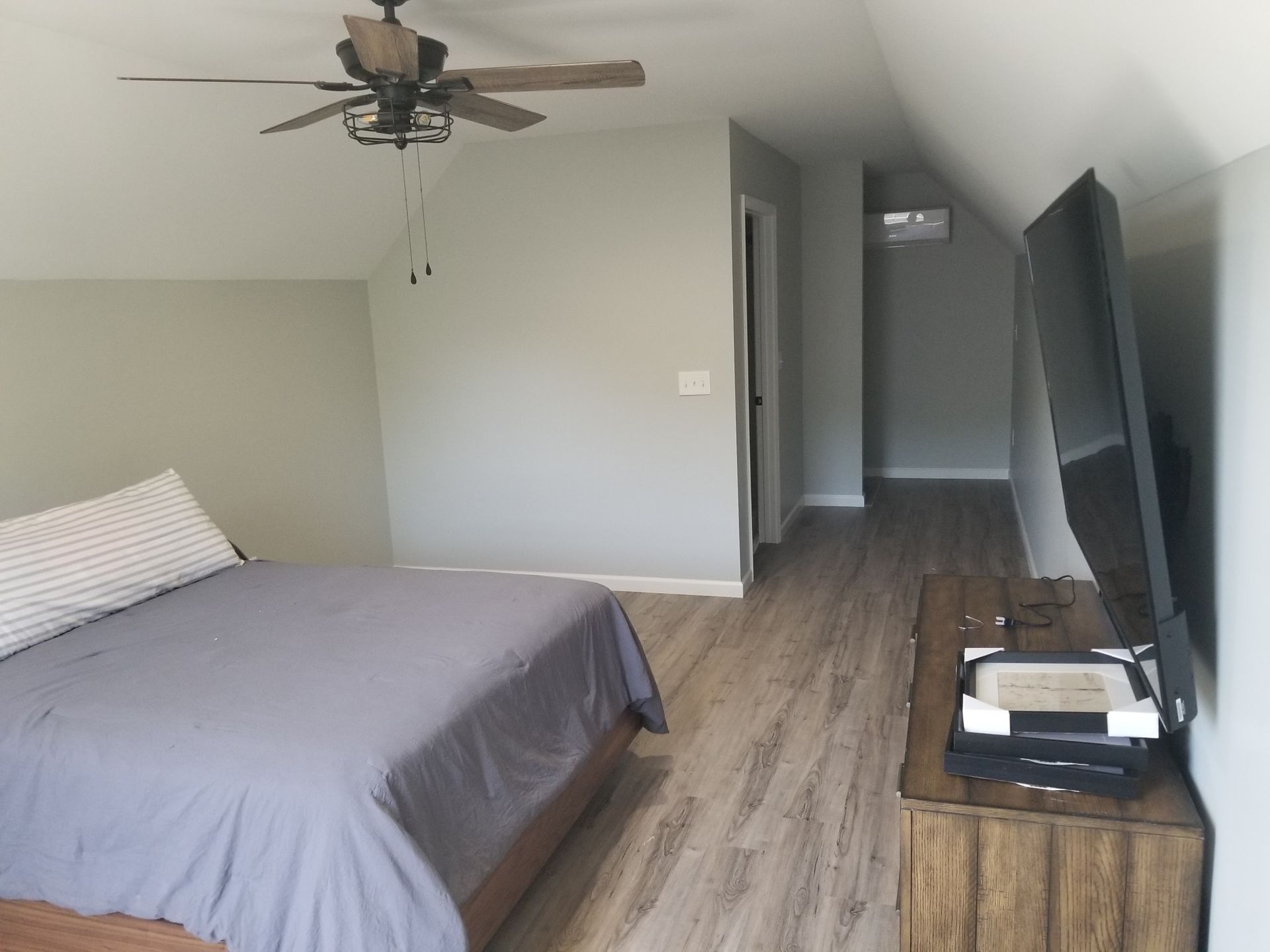 Bedroom with gray walls, wooden floor, bed, TV, ceiling fan, and doorway.
