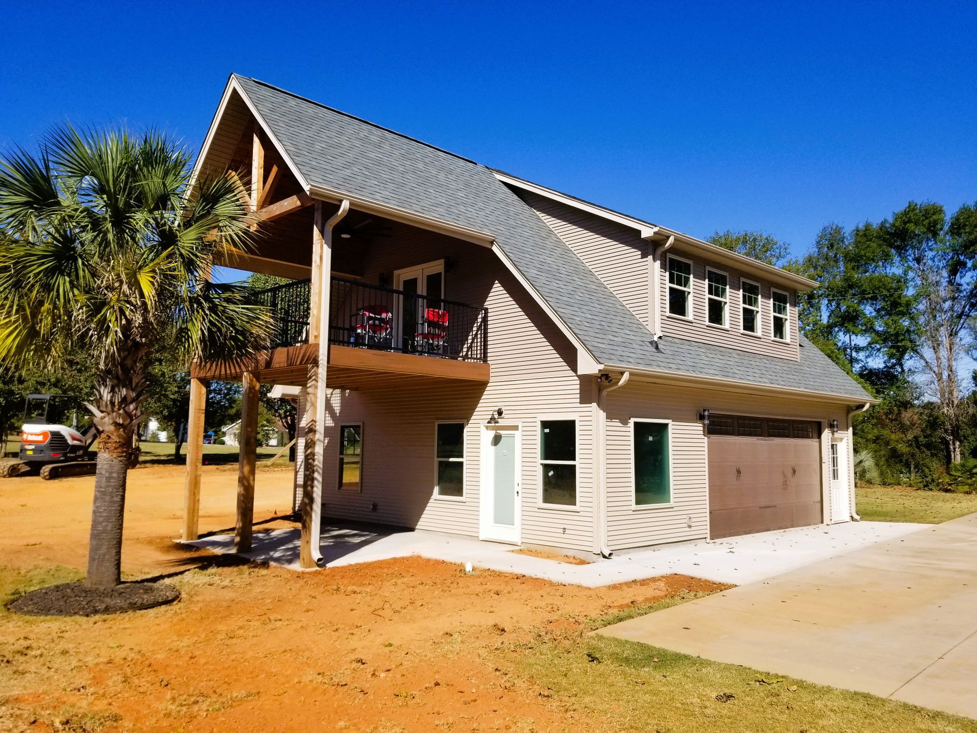 Two-story beige house with a balcony, gray roof, and a palm tree on a clear, sunny day.