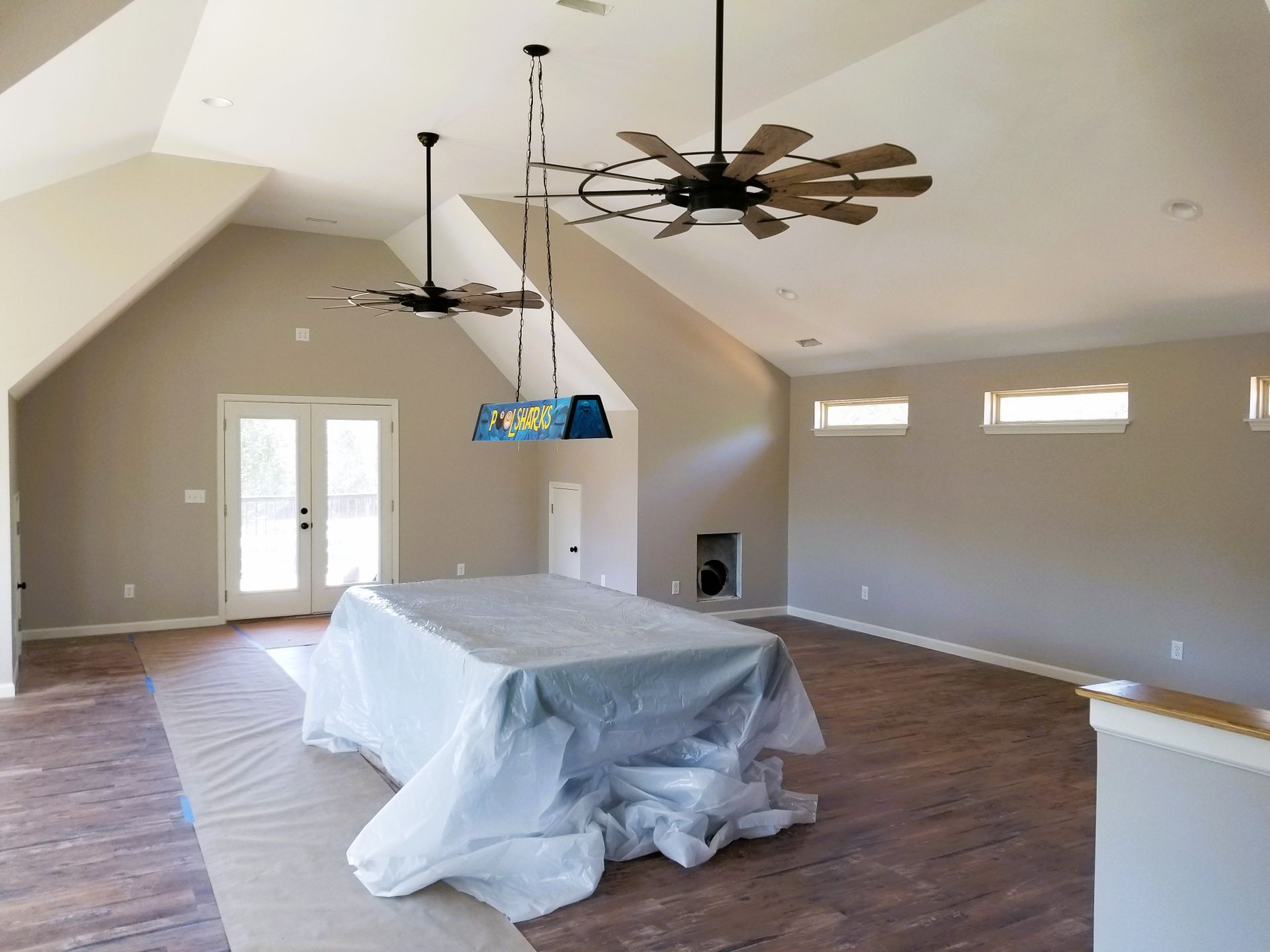 Spacious room with a covered pool table, two ceiling fans, and a chandelier; light-colored walls and wood-look flooring.